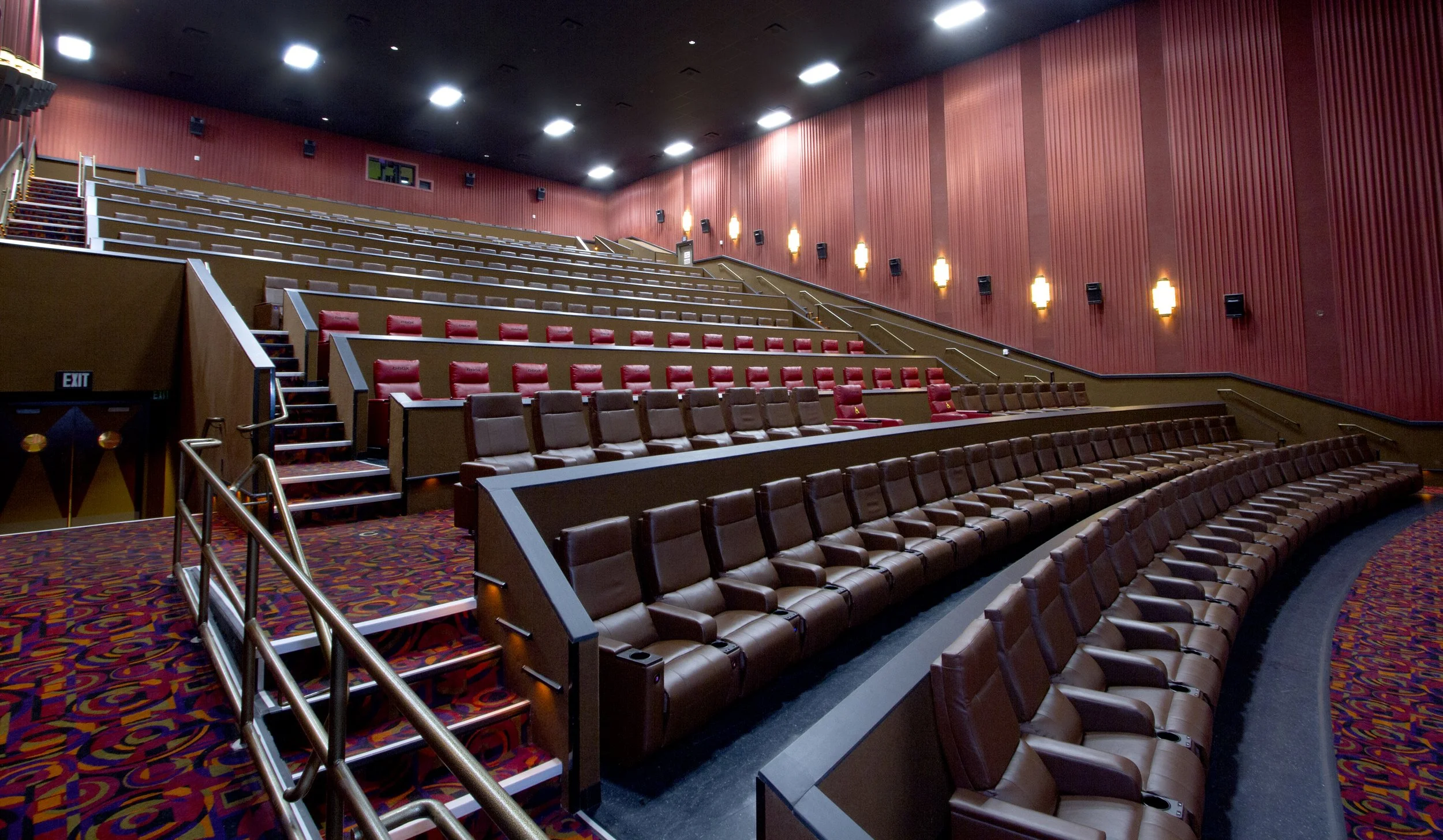 Inside a movie theater with rows of brown and red cushioned seats, red patterned carpet, and red walls with lighting fixtures.