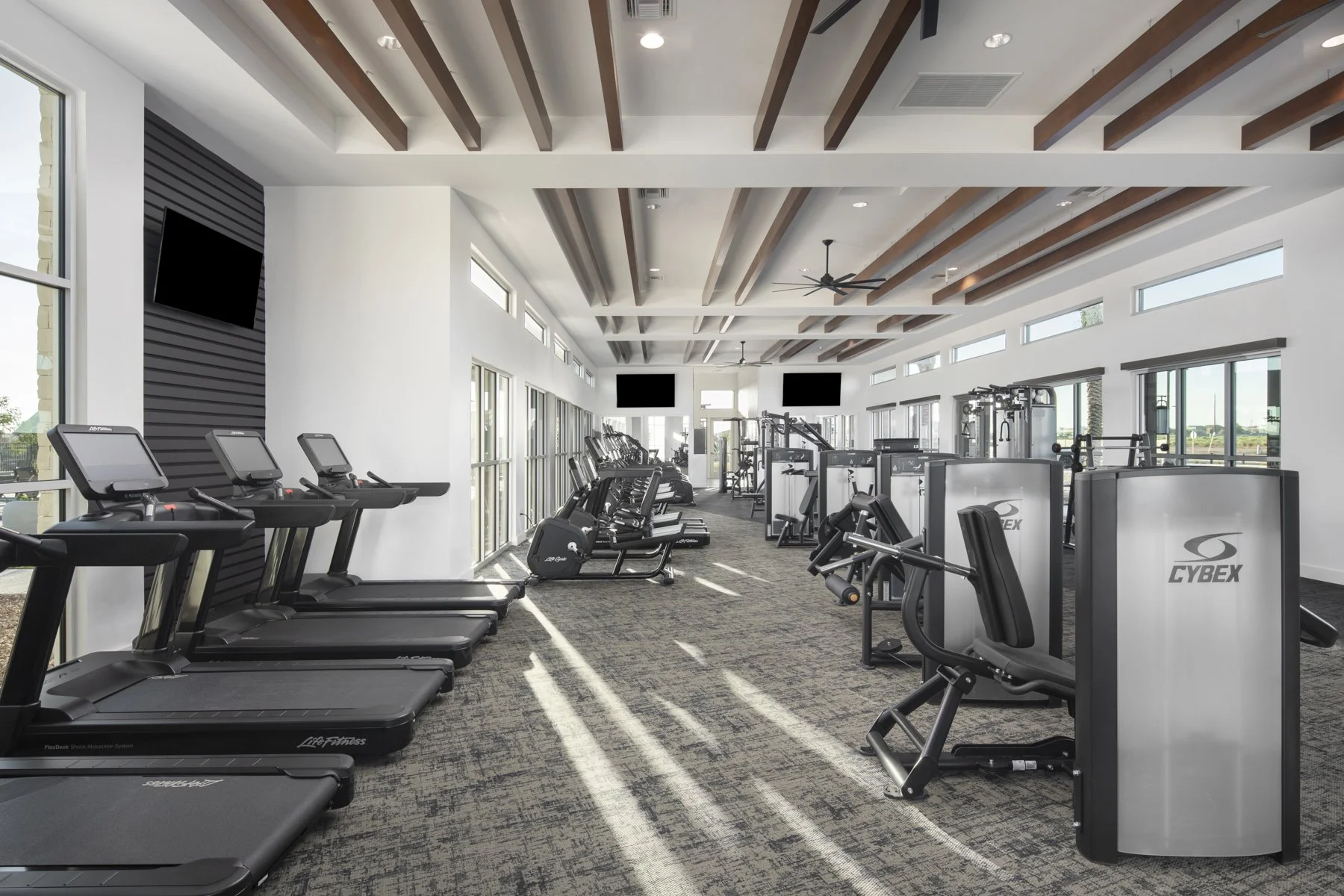 Modern gym with treadmills, cardio equipment, and weight machines, featuring large windows and wooden ceiling beams.