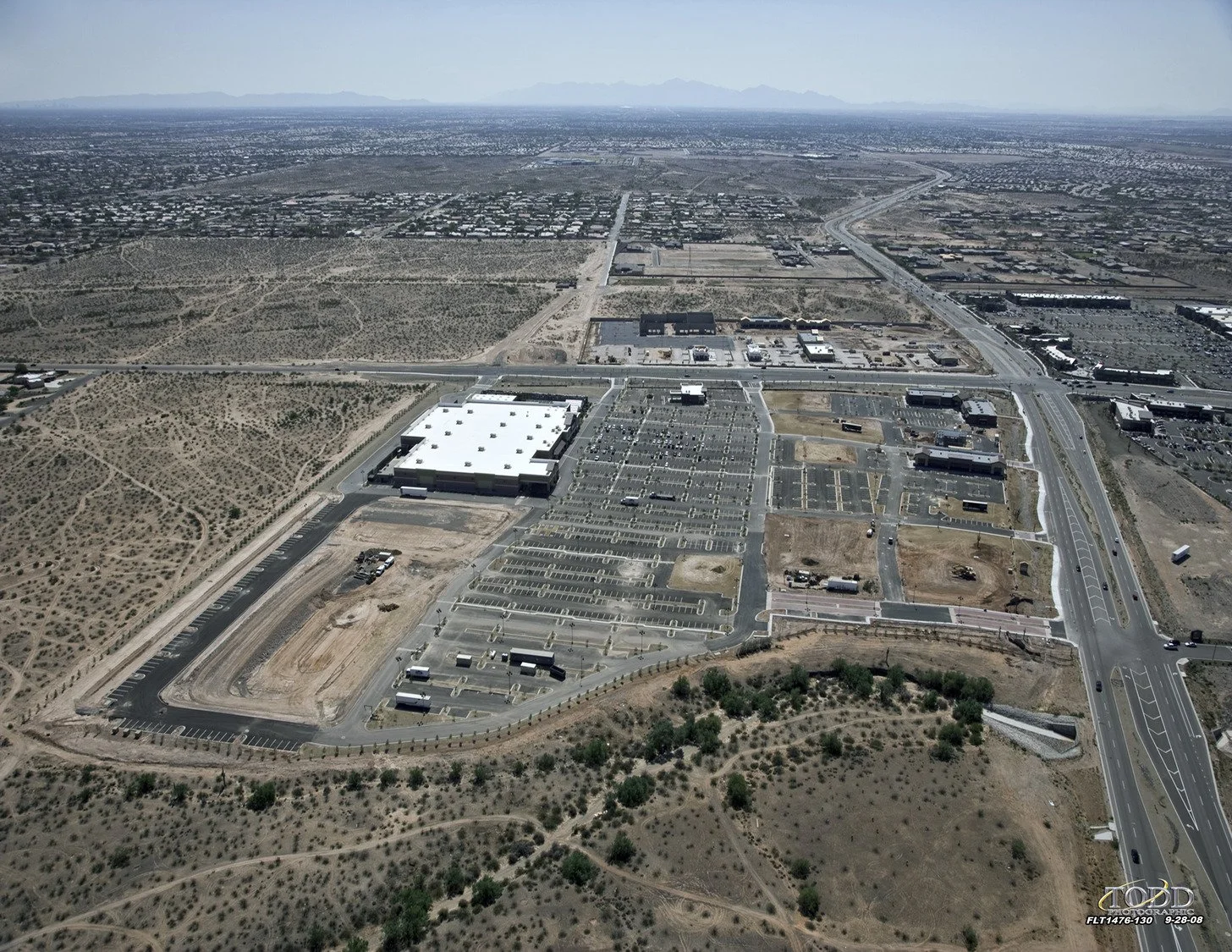 An aerial view of a large, mostly empty parking lot and commercial buildings in a desert landscape.