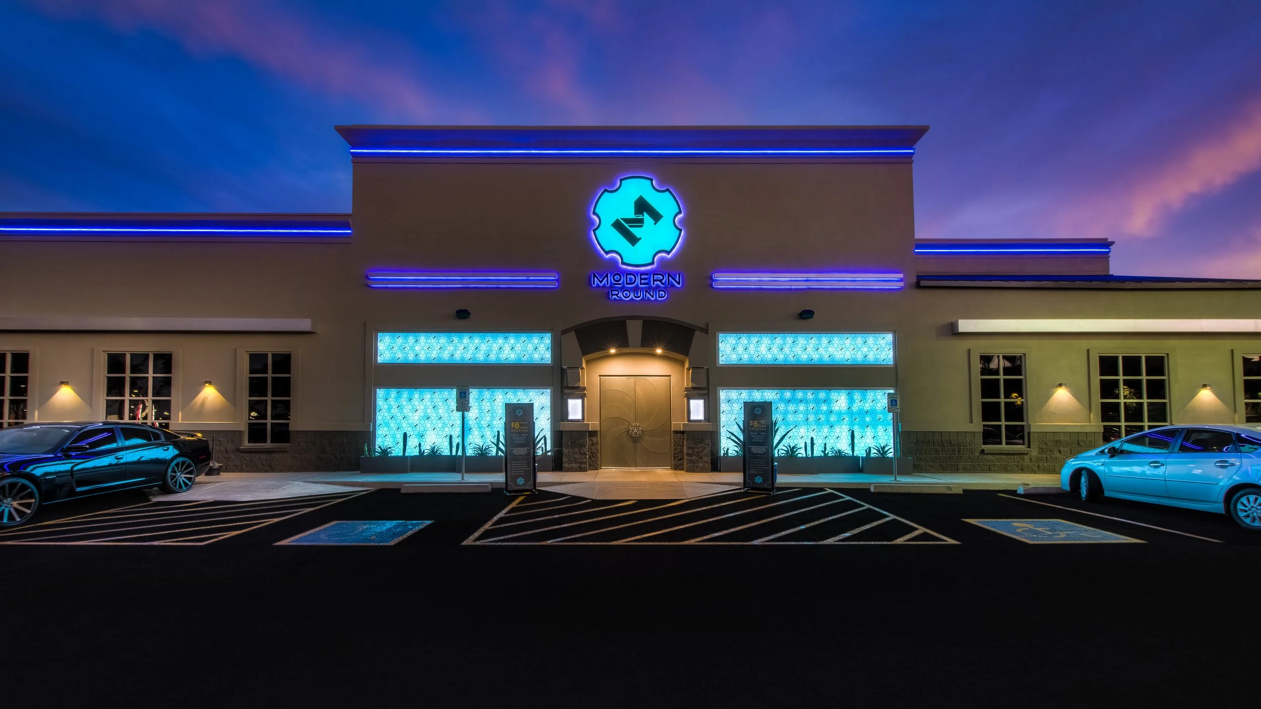 Night view of a modern building with bright blue neon lights and a sign that reads 'Modern Round'. The building has large windows with blue decorative lighting inside. There are cars parked in front and a parking lot with marked spaces, including a h