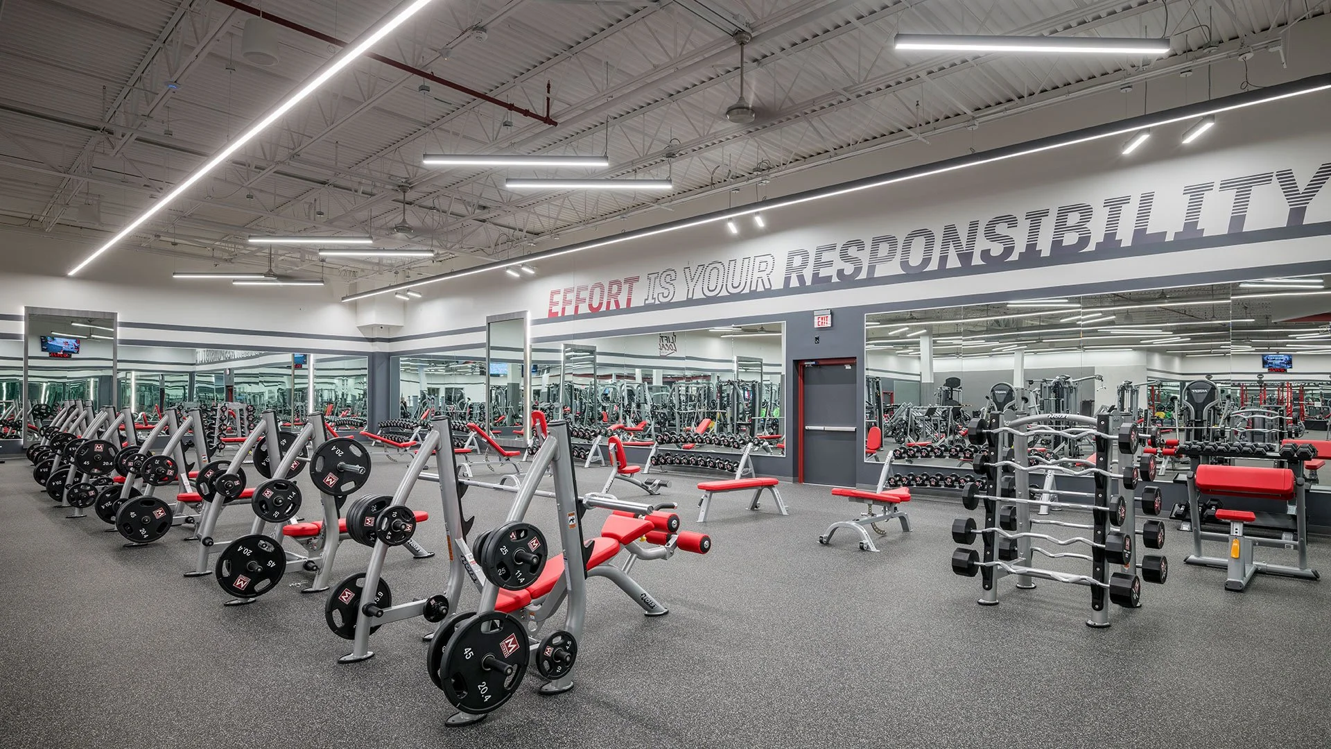 Empty gym with weightlifting benches, dumbbells, and various fitness equipment in a well-lit, modern space.