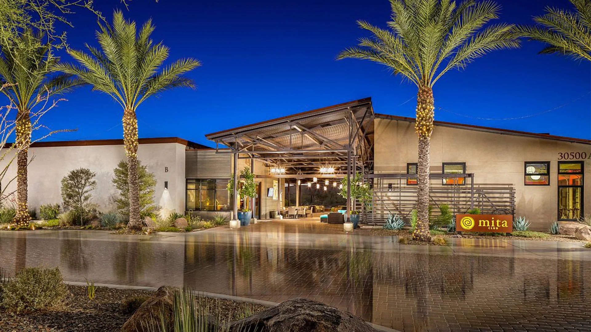 Exterior view of Mita Club during the evening, with a covered outdoor seating area, palm trees decorated with string lights, and landscape featuring desert plants and rocks.