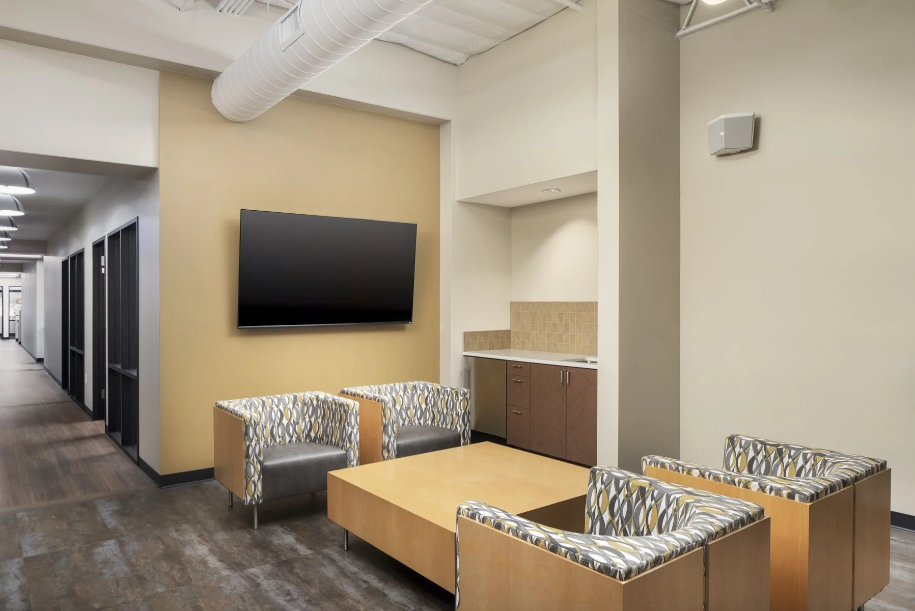 An empty waiting area with a large wall-mounted TV, four patterned chairs, a wooden coffee table, and a kitchenette with brown cabinets, beige backsplash, in a modern building corridor.