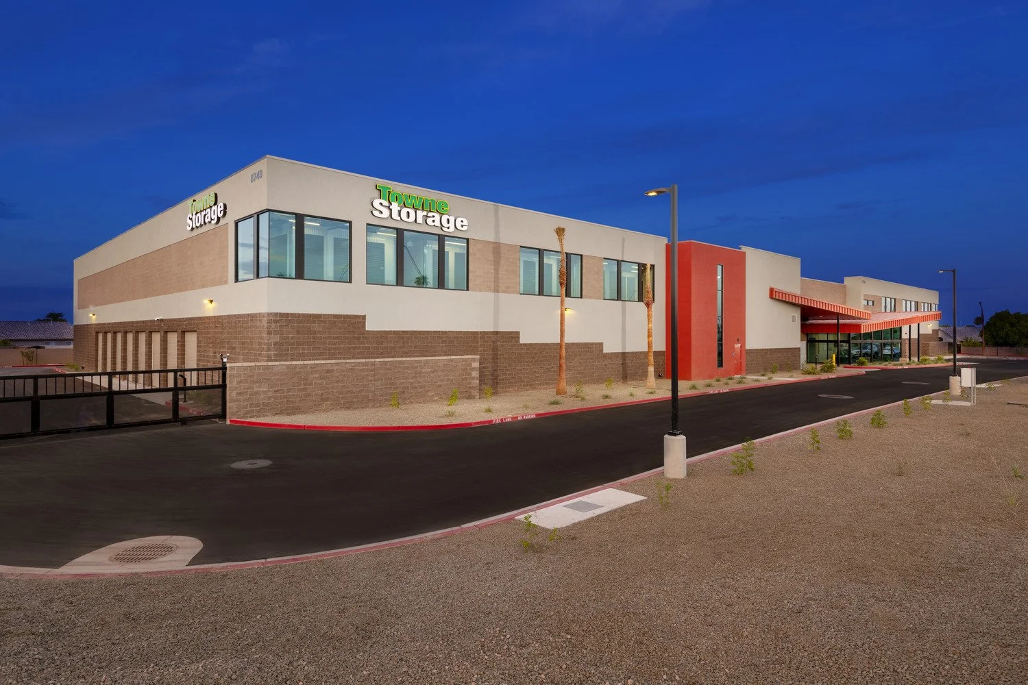 Exterior view of a modern storage facility building with a sign reading 'Towne Storage' and a smaller sign for 'Camo Storage,' taken during dusk under a darkening sky, with a paved parking area and streetlights.