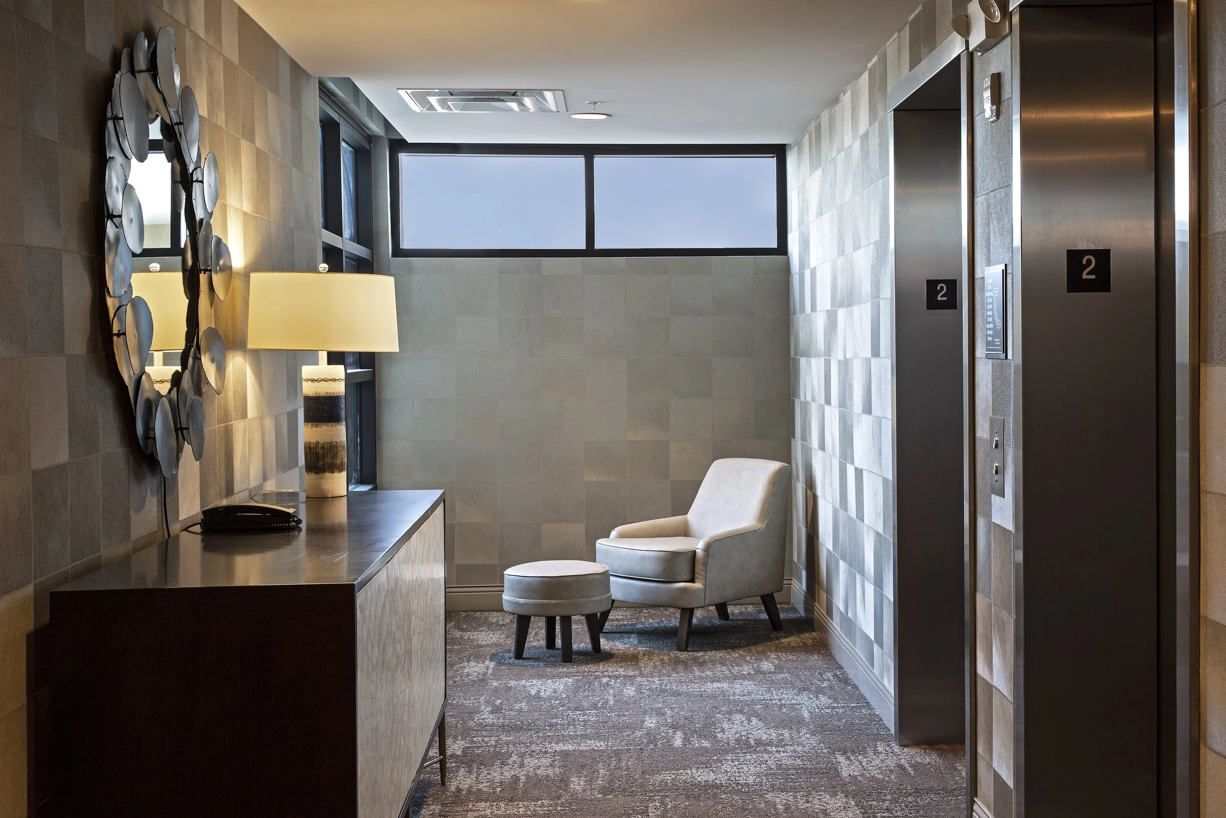 An elevator lobby with two elevators on the right, a beige armchair and matching ottoman, a wooden console table with a lamp and a decorative mirror, a telephone, and a window with blue sky visible outside.