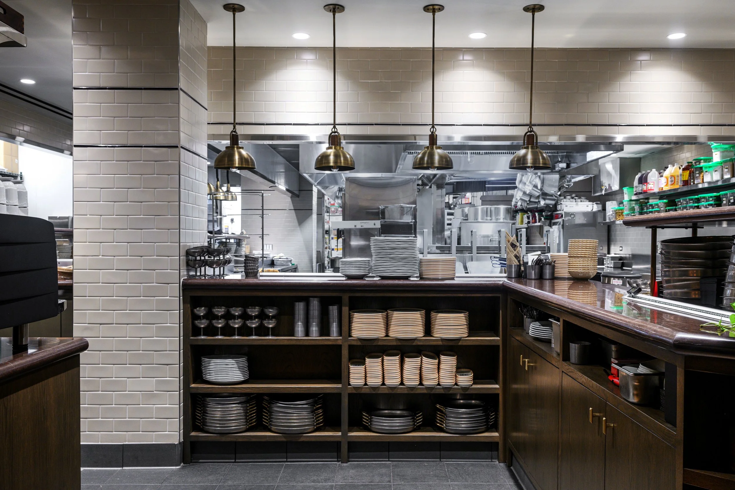 View of a restaurant kitchen with a bar counter in the foreground, shelves filled with plates and bowls, and industrial kitchen appliances in the background.