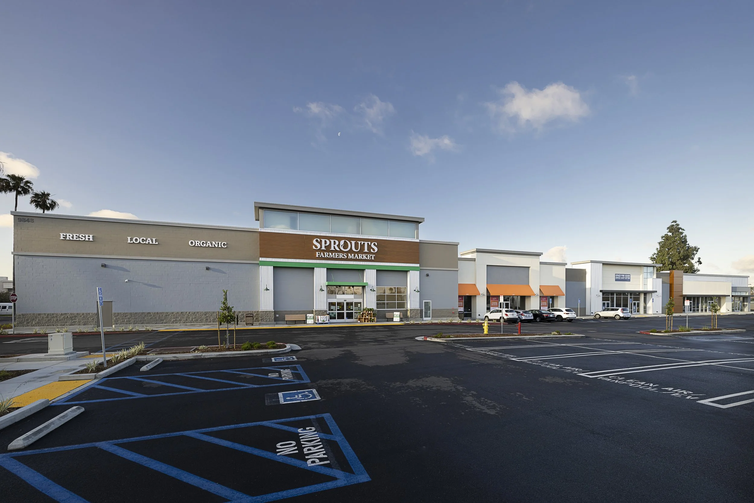 Parking lot in front of Sprouts Farmers Market with parking spaces marked disabled and no parking signs, and a grocery store building with large windows, a sign that says 'Fresh Local Organic,' and orange awnings, under a clear blue sky with a few cl