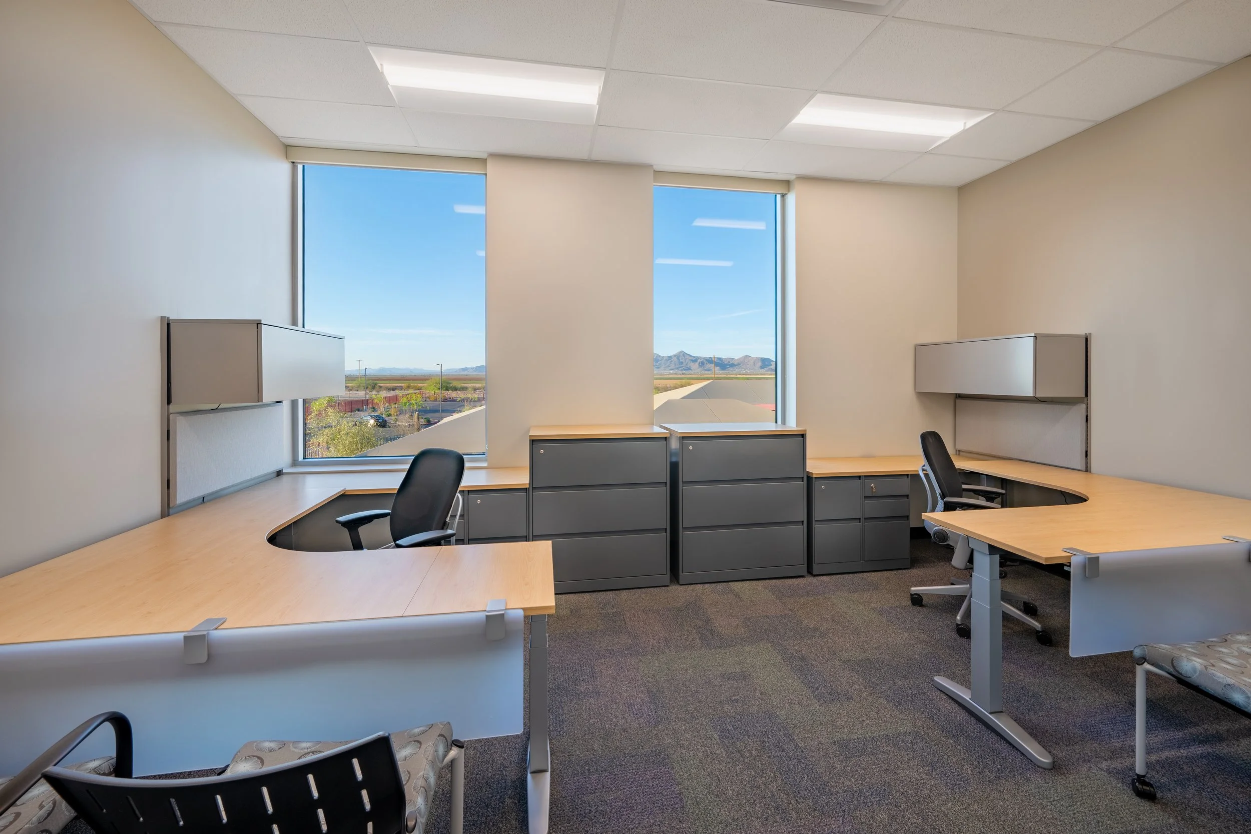 Empty office with L-shaped wooden desks, black office chairs, gray filing cabinets, and large windows showing a mountain landscape and blue sky.