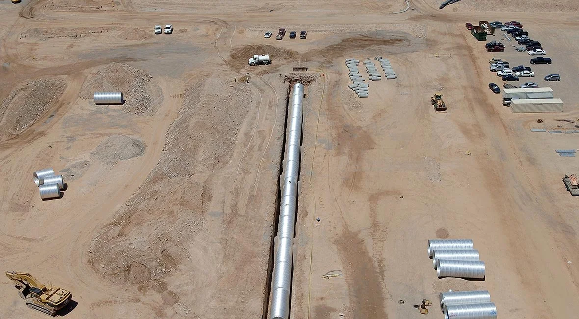 Aerial view of a construction site with a long pipe running through the middle, construction vehicles, parked cars, and various equipment scattered around.
