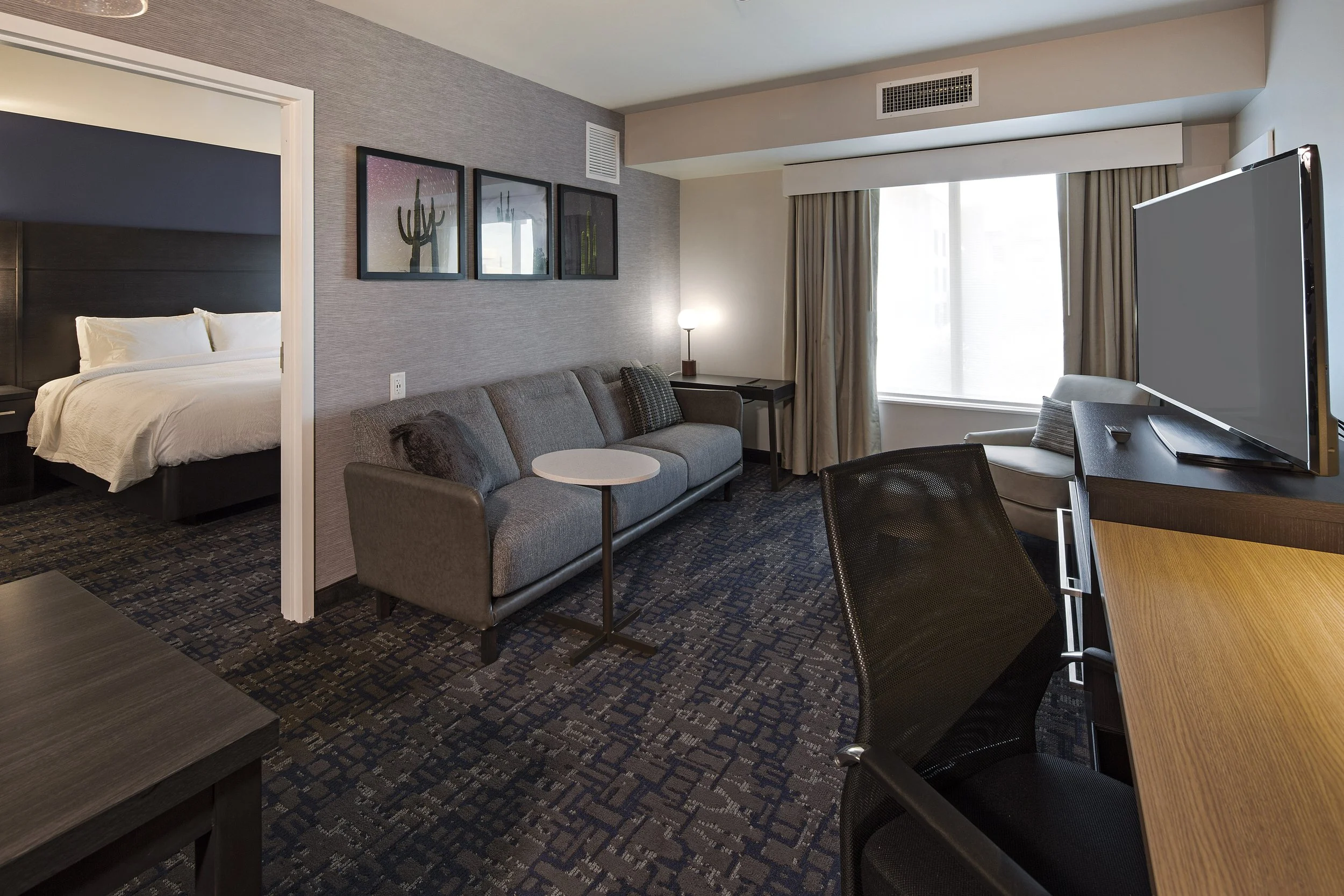 Room with a gray couch, three framed cactus pictures on the wall, a dark wood desk with a lamp, a large window with beige curtains, a TV on a black stand, and part of a bed visible through an open door.