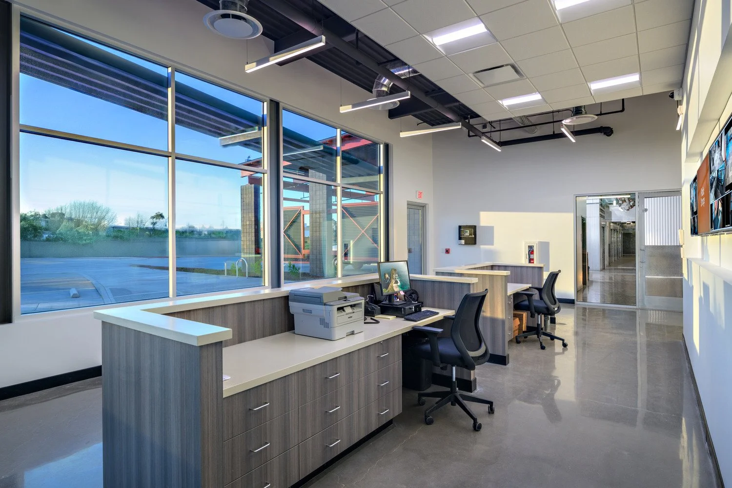 Empty modern office reception or waiting area with large windows, desks, chairs, and office equipment.