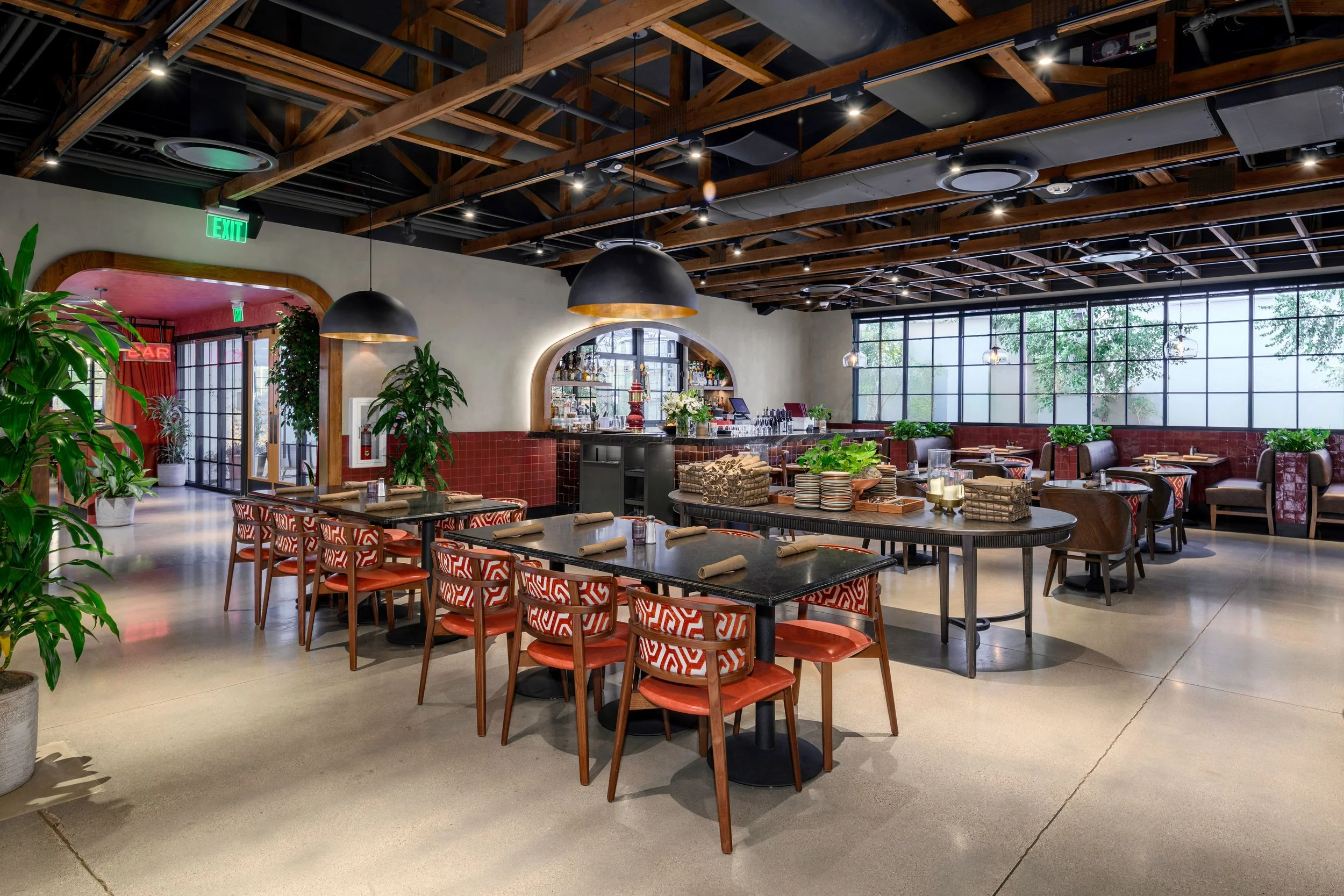 Interior of a modern restaurant with tables and chairs, a bar with bottles and glasses, large windows bringing in natural light, and decorative plants.