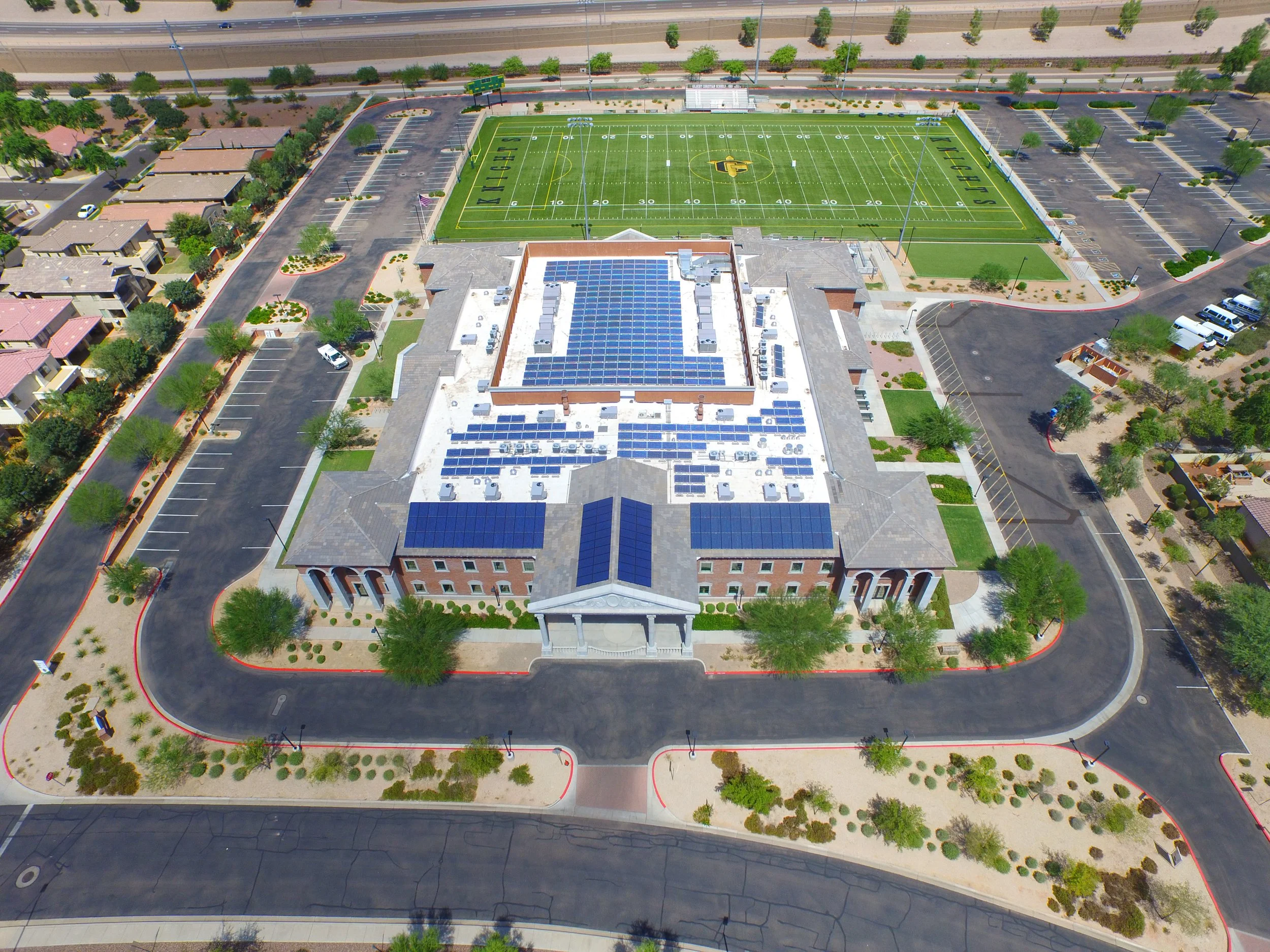 Aerial view of a school campus with a building that has solar panels, a large parking lot, and a football field in the background.