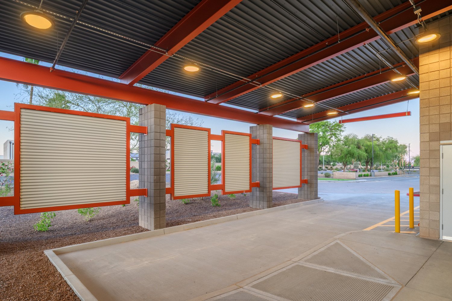 Empty drive-thru area with shuttered service windows, concrete pavement, and a landscaped area with small plants and trees outside.
