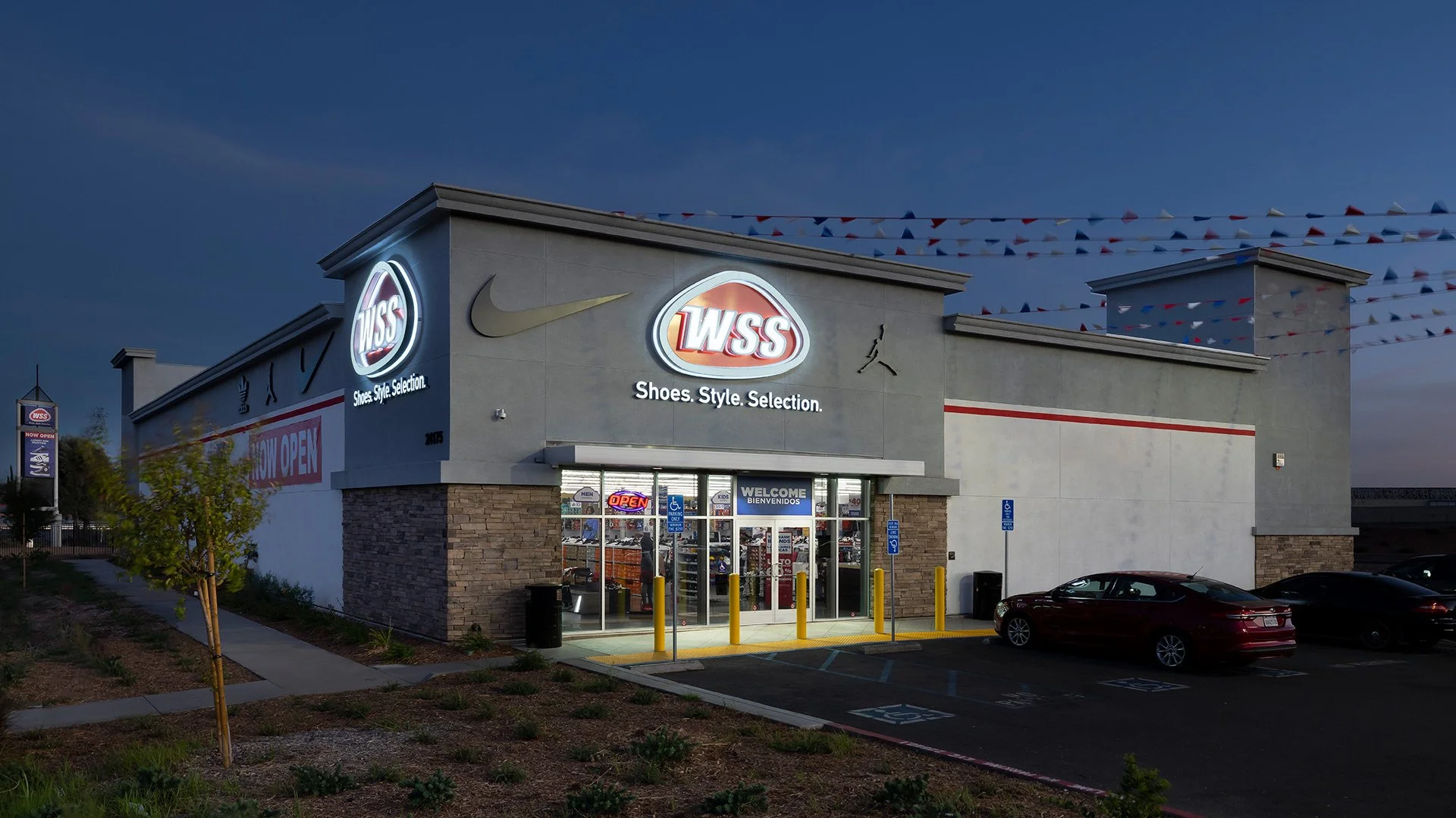Exterior of a WSS shoe store with illuminated signage, parking lot with a few cars, and a sidewalk in the evening.