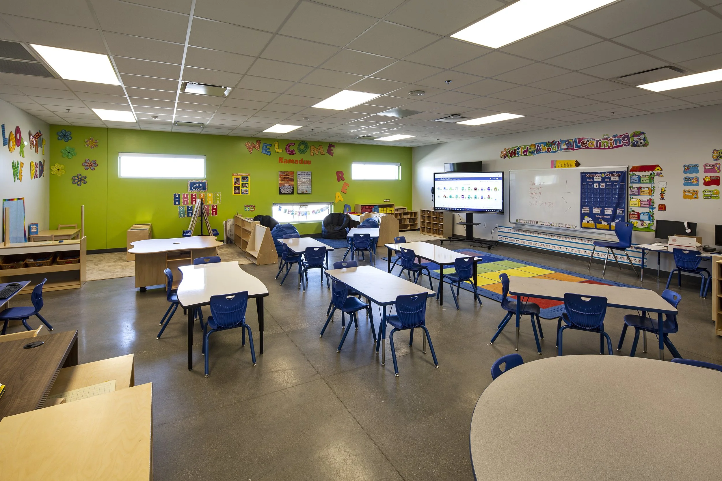 Empty kindergarten classroom with colorful decorations, small tables and blue chairs, educational posters, a large digital display, and a whiteboard.