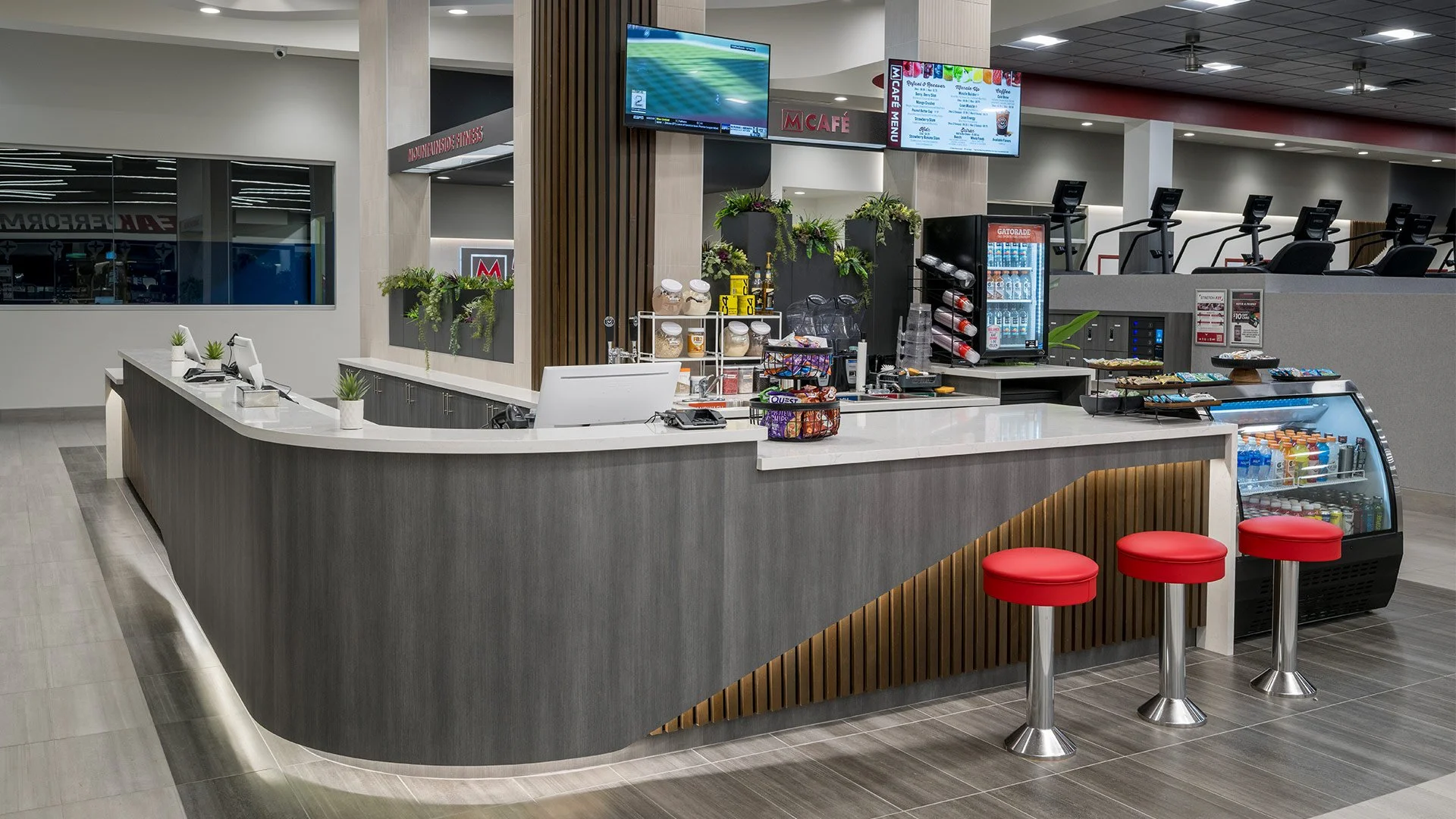 Cafe counter with three red stools, refrigerated drinks, snacks, and a touchscreen register in a modern indoor setting.