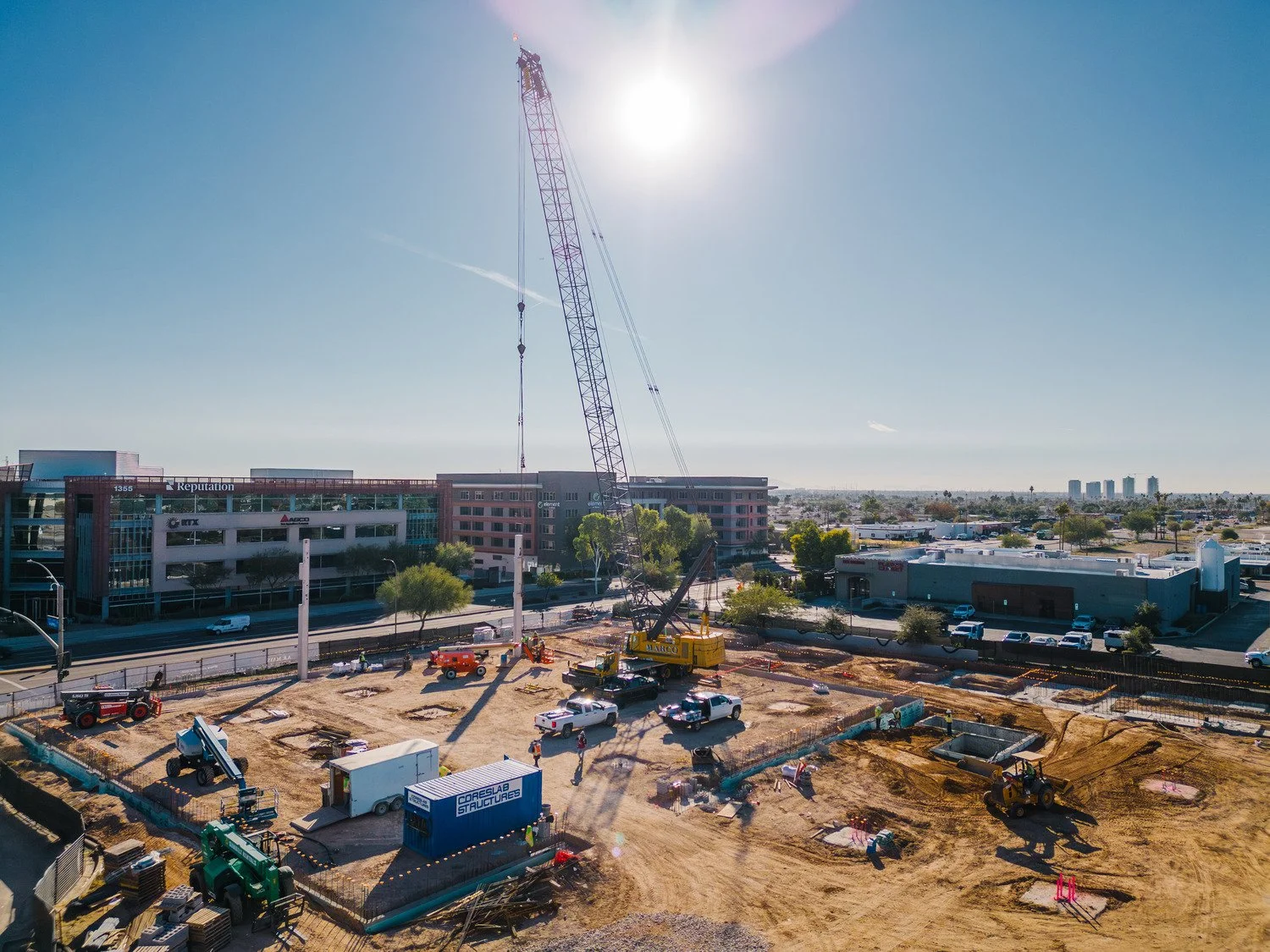 Construction site with a large crane, construction vehicles, and buildings in the background on a sunny day.