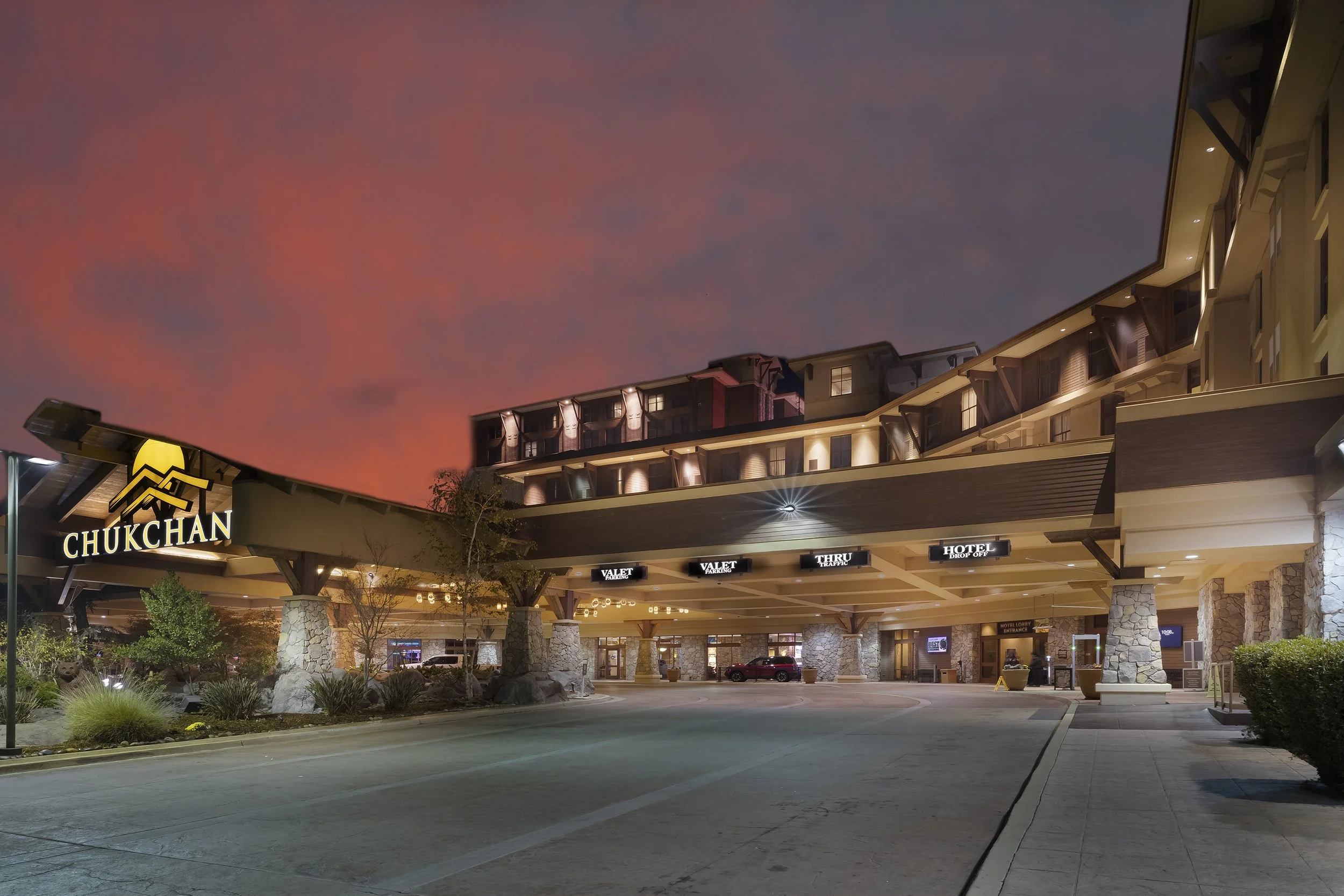 Exterior view of the Chukchan hotel at dusk with a colorful sunset sky, illuminated signs, and a parking area in the foreground.