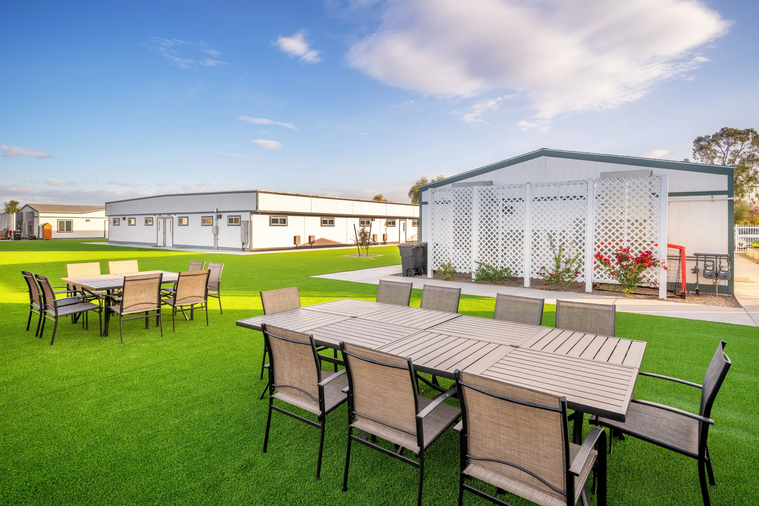 Outdoor patio area with two sets of patio tables and chairs on well-maintained green grass, with white buildings and a partly cloudy sky in the background.