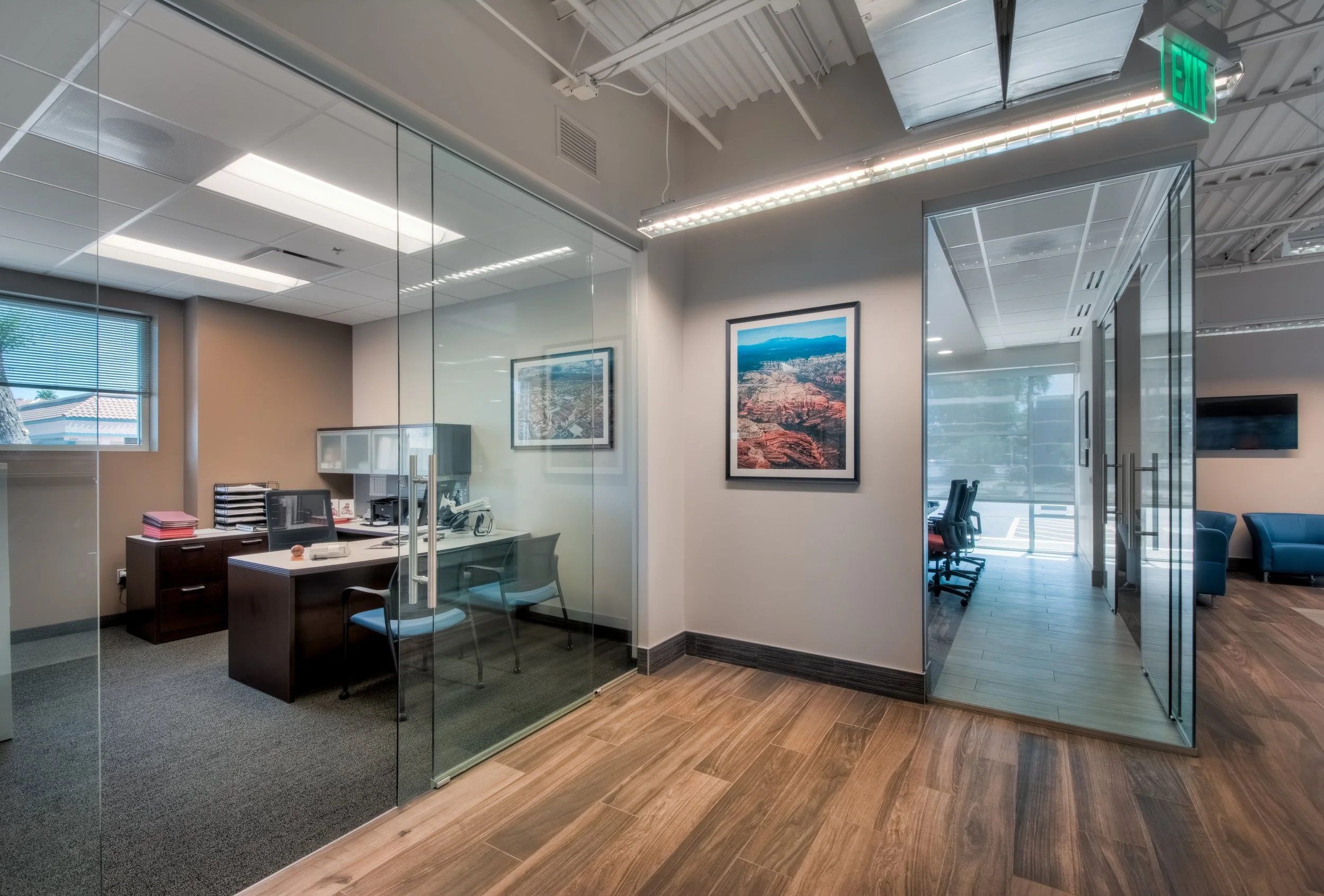 Modern office with glass walls, framed pictures, conference room with chairs, and wooden flooring.
