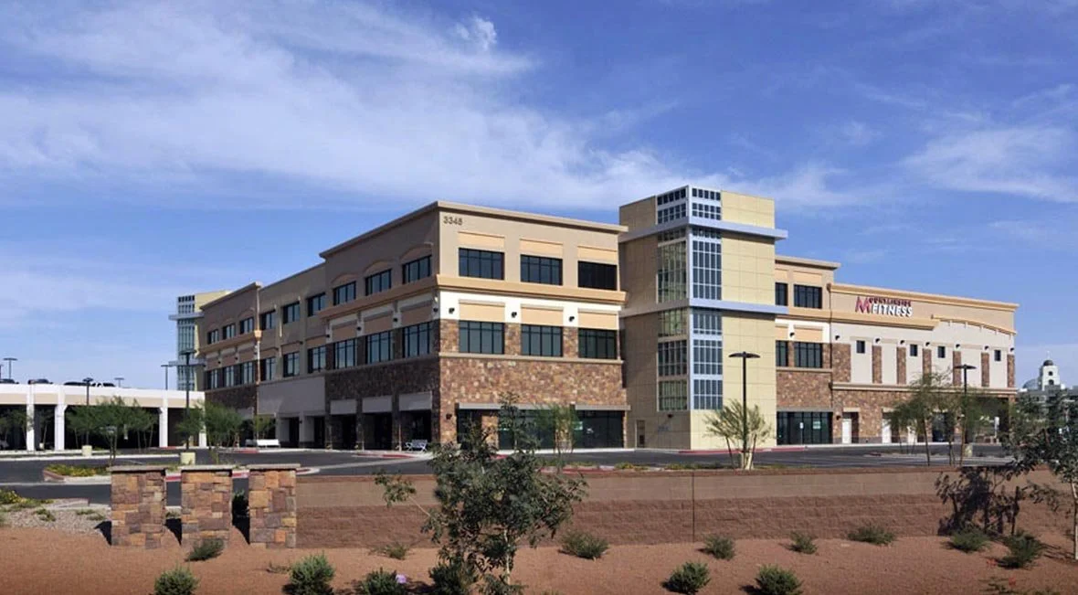 A multi-story commercial building with a modern design, featuring large windows, a mix of stone and beige siding, and a sign indicating a fitness center. The building is surrounded by a landscaped area with small bushes and trees, under a partly clou