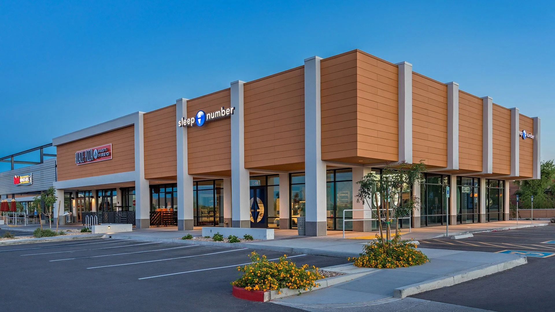 Empty shopping plaza with a Sleep Number store and Blaze Pizza restaurant, parking spaces, flowers, and a clear blue sky.