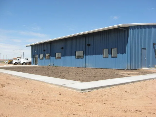 A new blue industrial building with a sloped roof and multiple small windows, with a concrete sidewalk and dirt area in the foreground, and a few white trucks parked in the background.