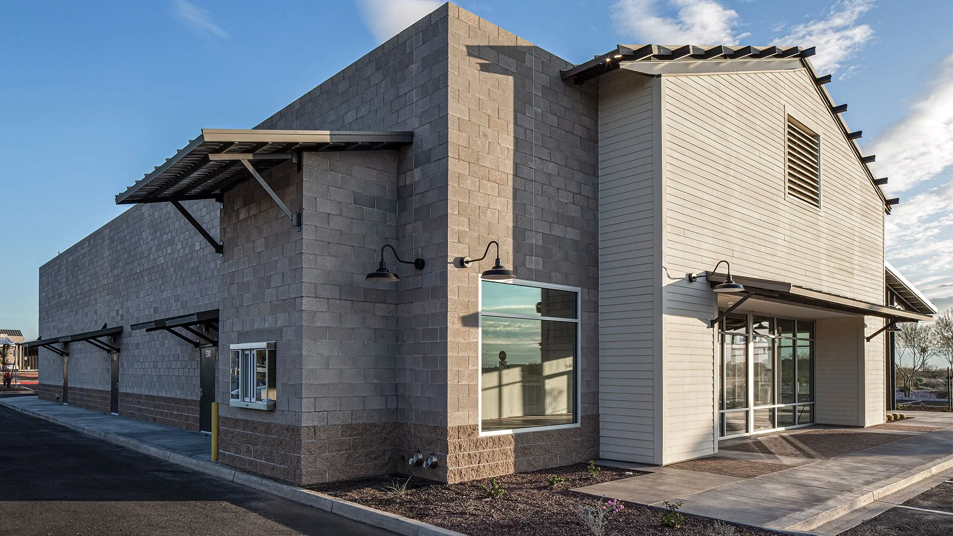 Modern commercial building with beige and gray exterior, large glass entrance doors, windows, and outdoor lighting fixtures.