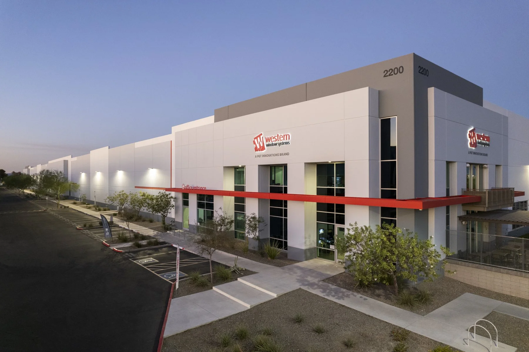 Exterior view of a modern commercial building with the sign 'Western Window Systems'. The building has large windows, a parking lot with designated spots, some small trees, and exterior lighting.