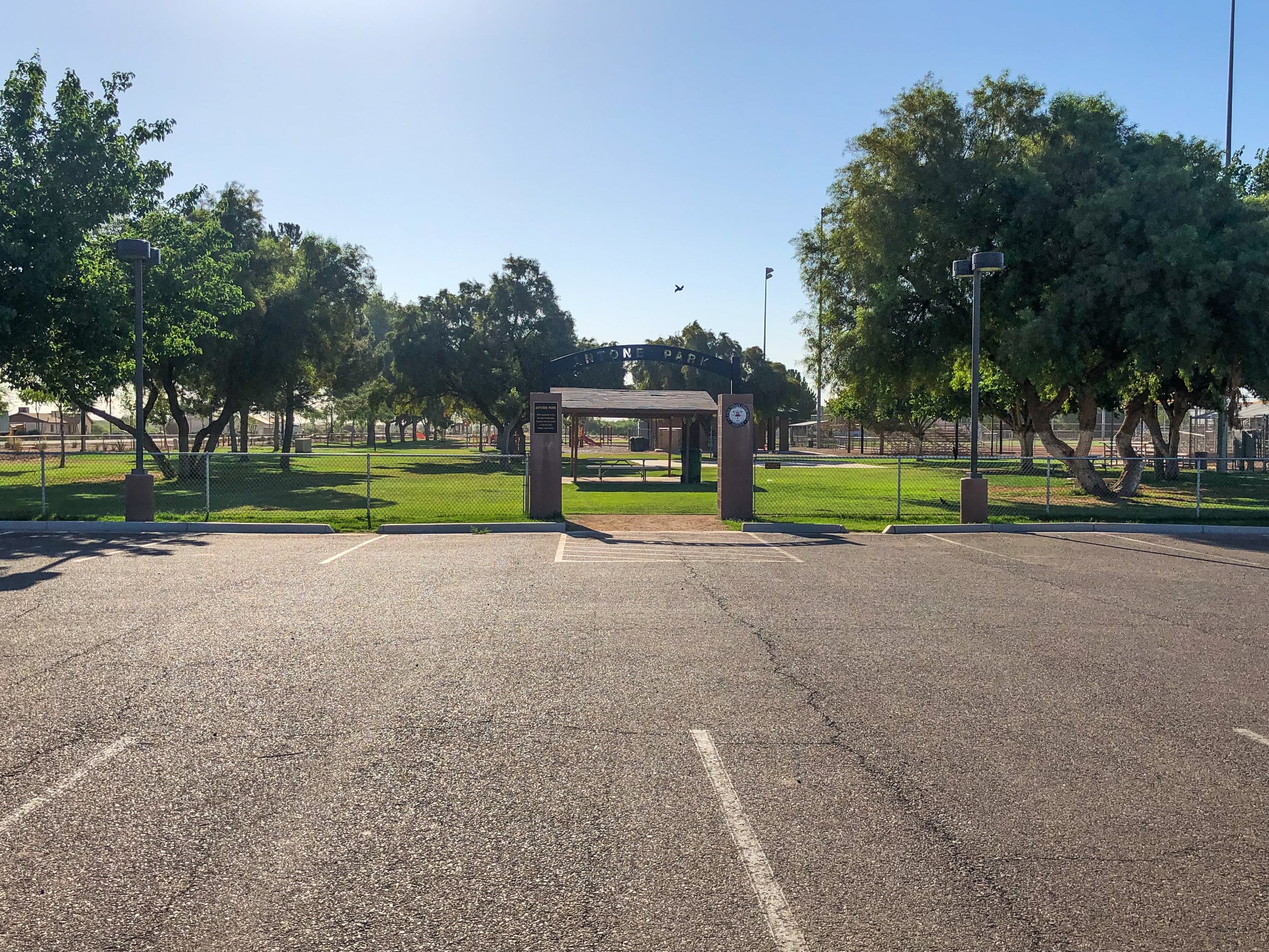 View of a park entrance with a sign reading 'Bite One Park,' surrounded by green grass, trees, and some picnic tables underneath a shelter. The parking lot in the foreground is mostly empty, with a few marked parking spaces, and the sky is clear and 