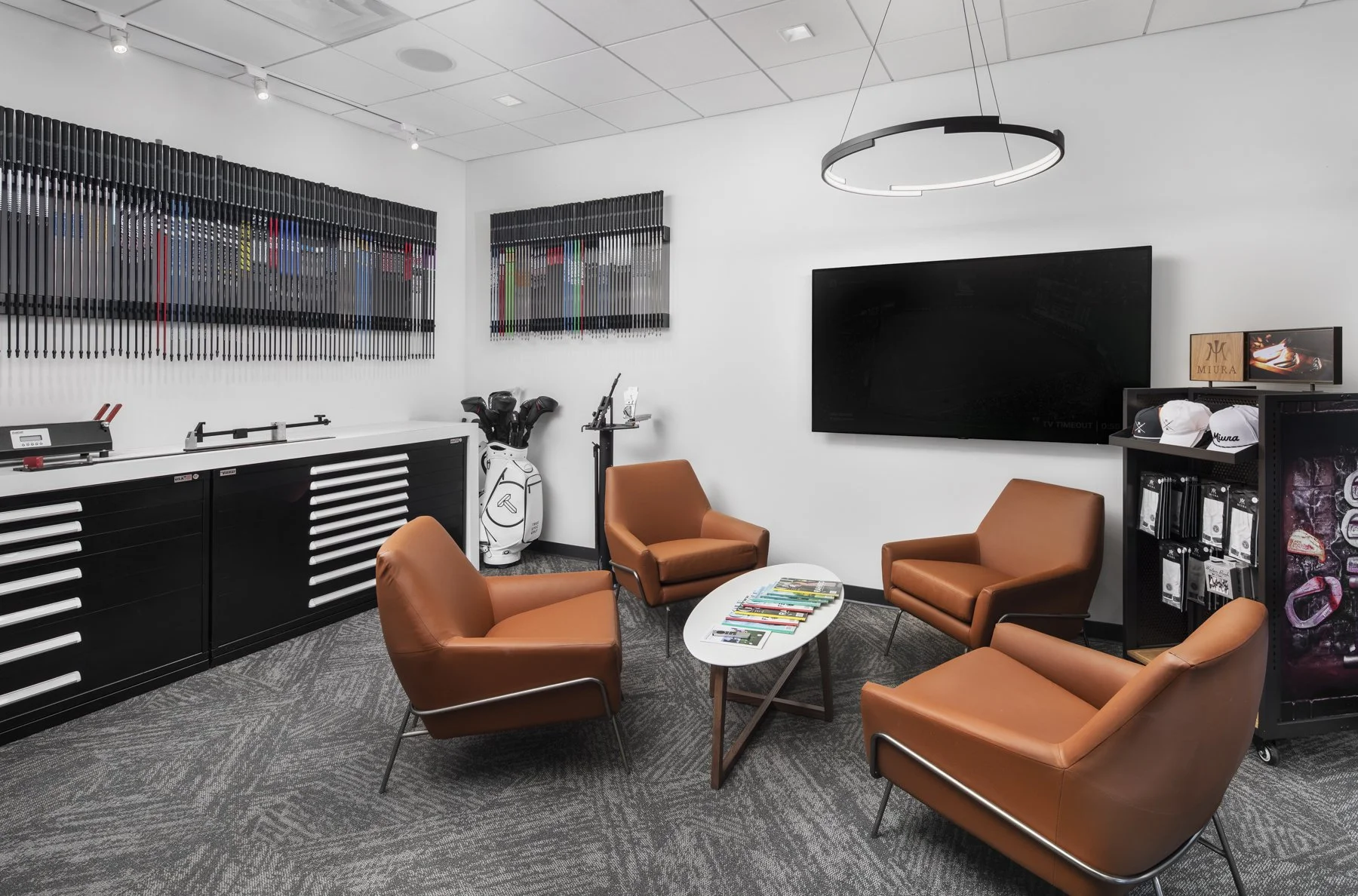 A modern golf club lounge with four brown leather chairs arranged around a white coffee table with magazines, a large flat-screen TV on the white wall, golf clubs in a bag, and golf-themed artwork on the wall.