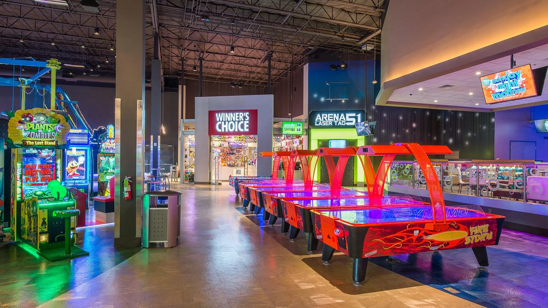 Indoor arcade with multiple electronic games, including air hockey tables with red and black designs, surrounded by seating areas and brightly lit gaming machines.