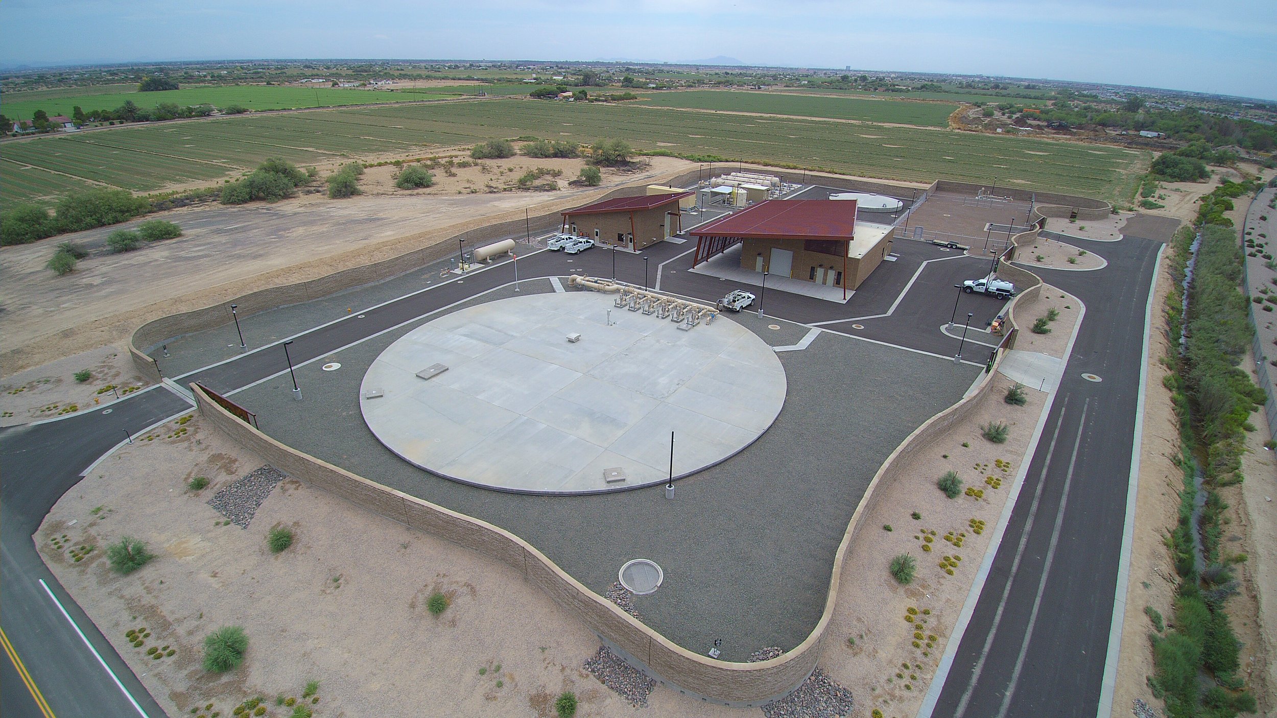 Aerial view of a modern building complex with a large flat roof, surrounded by a landscaped yard, parking lot, and roads, in a semi-arid area with fields and farmland in the background.