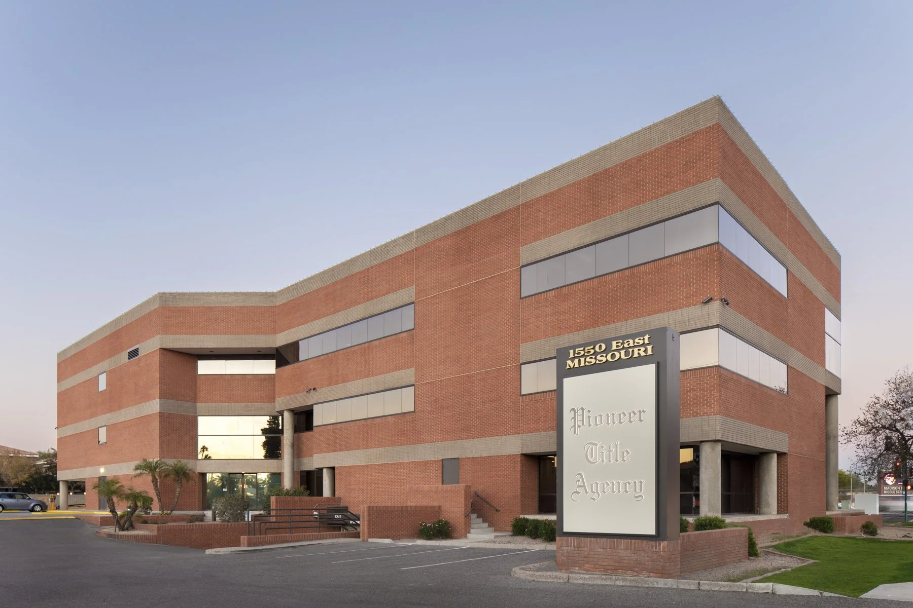 A three-story red brick office building with horizontal bands of beige and several large tinted windows; a sign reads '1550 East Missouri Pioneer Title Agency'; landscaped area with small palm trees in the foreground.