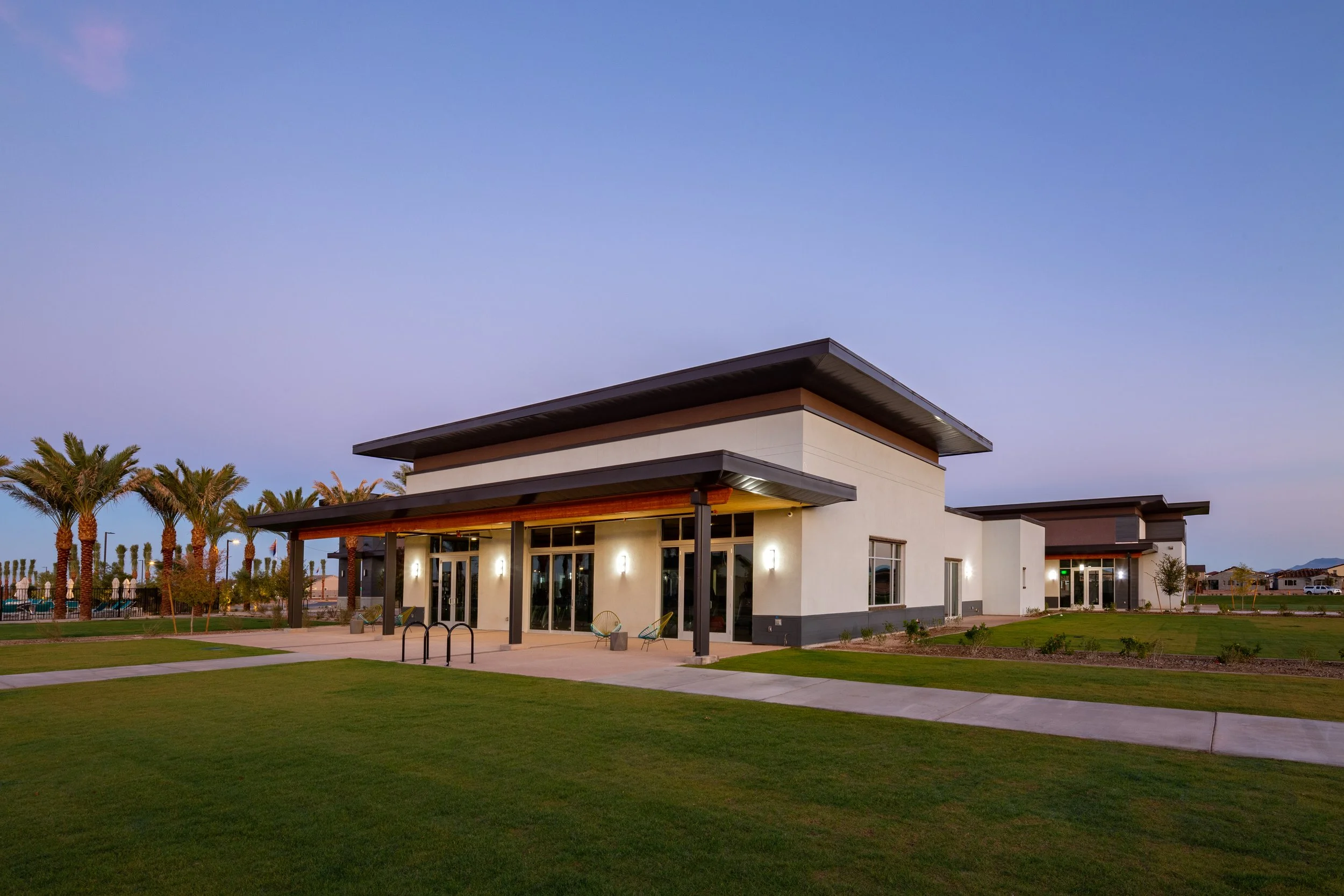 Modern building with large windows, outdoor seating, surrounded by green lawns and palm trees, under a evening sky.