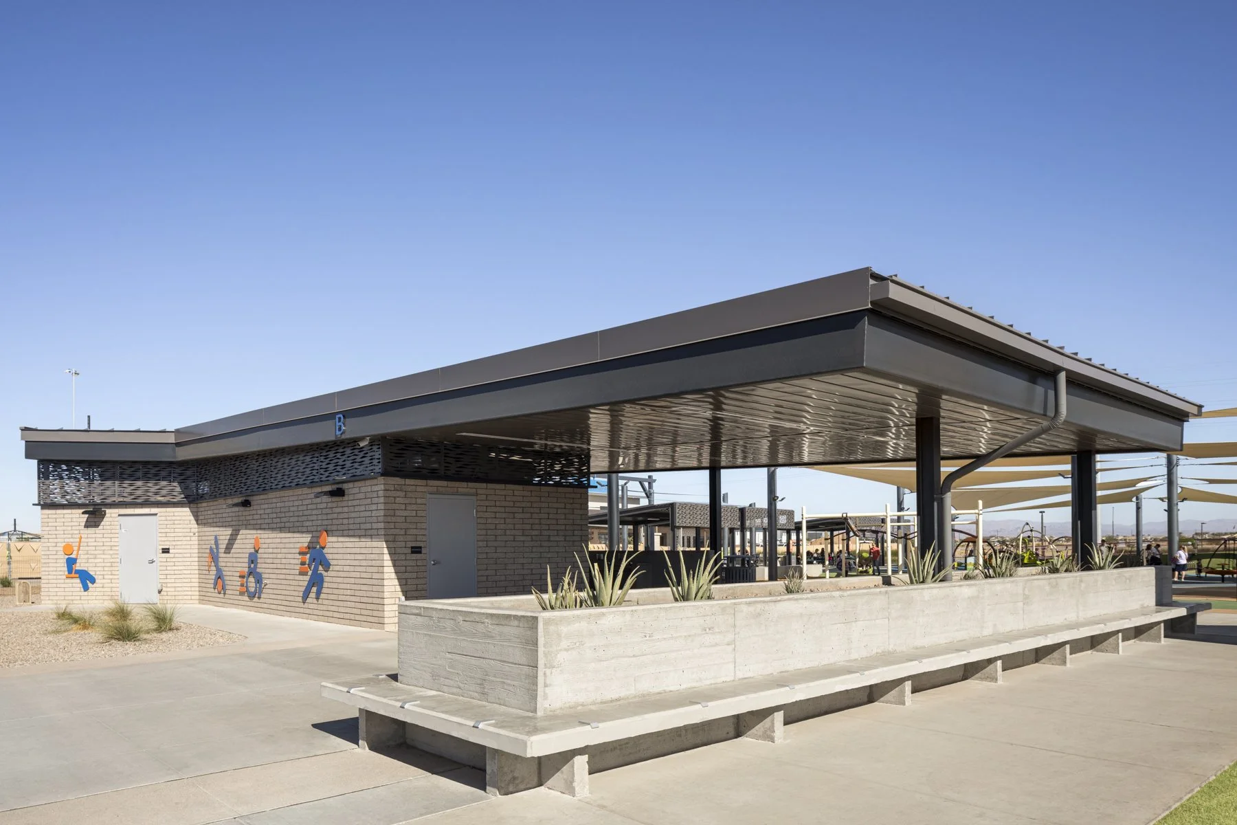 Public restroom facility with a brick and metal exterior, restroom sign painted on the wall, and an open-air shaded waiting area with seating and plants.