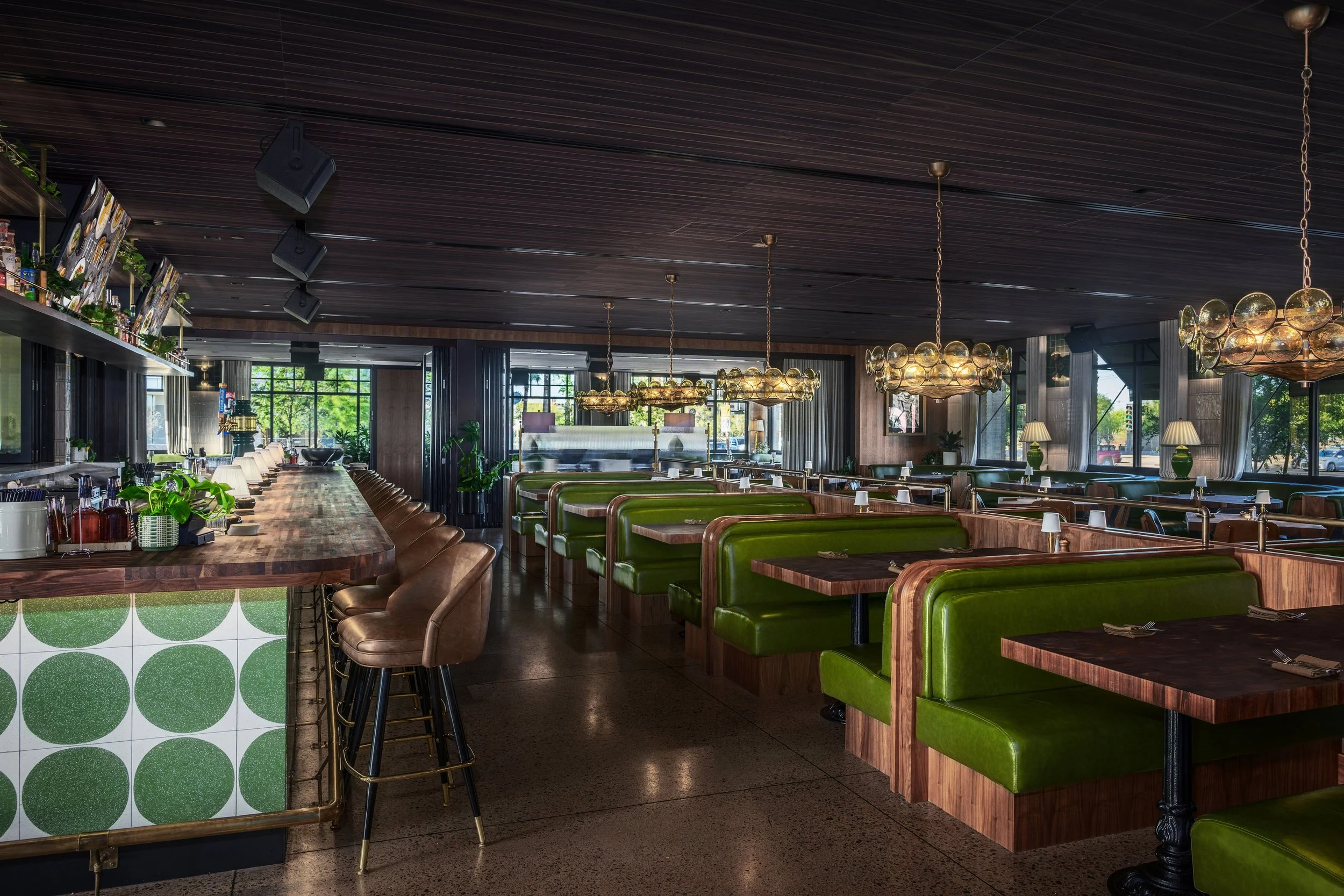 Empty restaurant interior with green booths, dark wood tables, bar seating area, large windows, hanging chandeliers, and greenery outside
