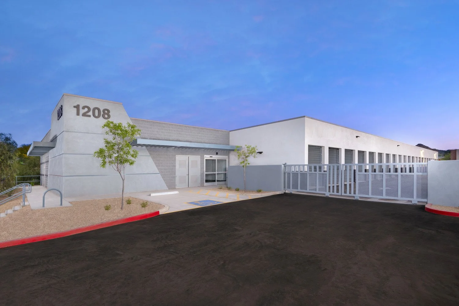 Modern building with white walls, marked with the number 1208, surrounded by a parking lot with a handicapped parking space, small trees, and a clear evening sky.