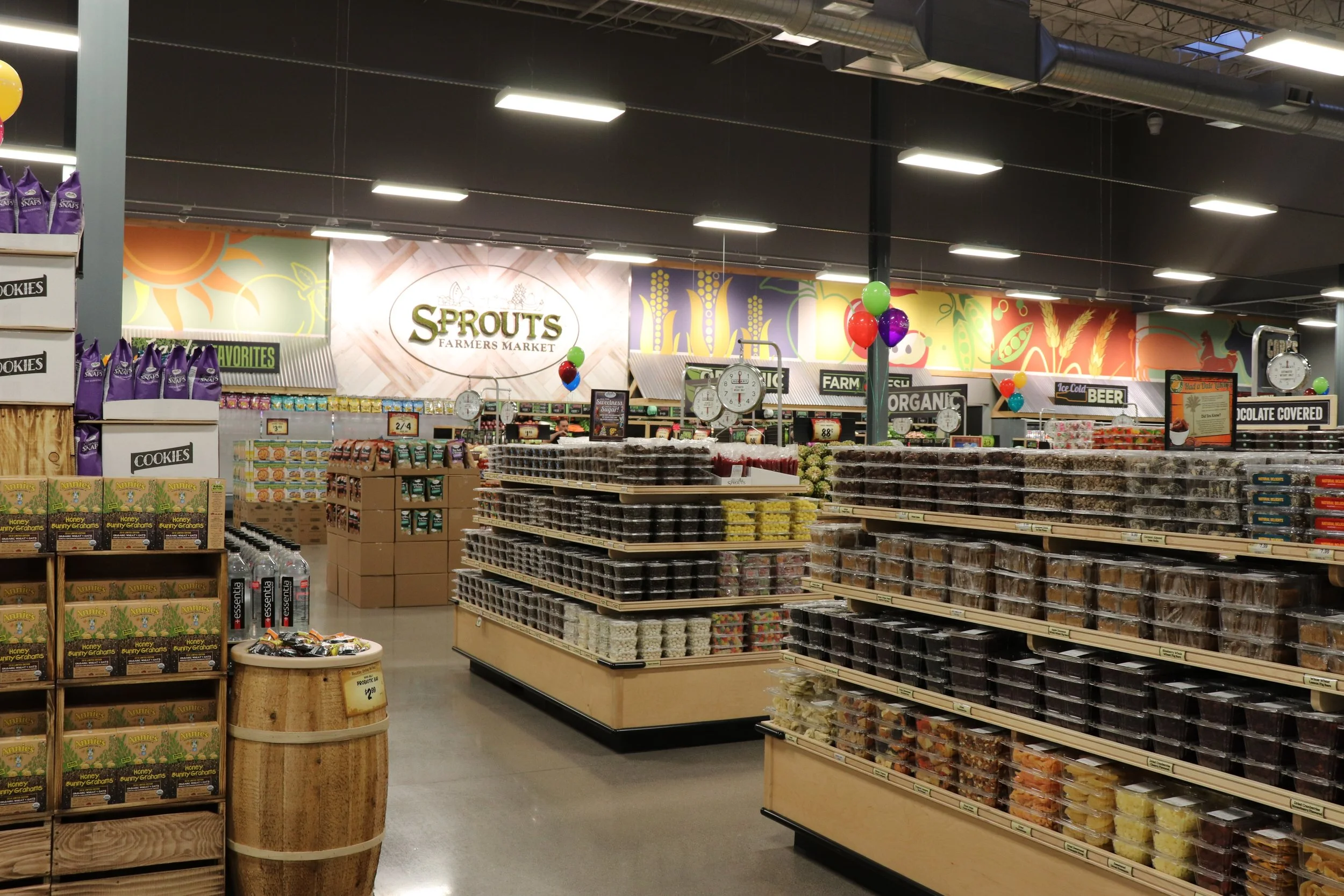 Interior of a grocery store with various shelves stocked with baked goods, candies, and snacks, and a sign that reads 'Sprouts Farmers Market' in the background.