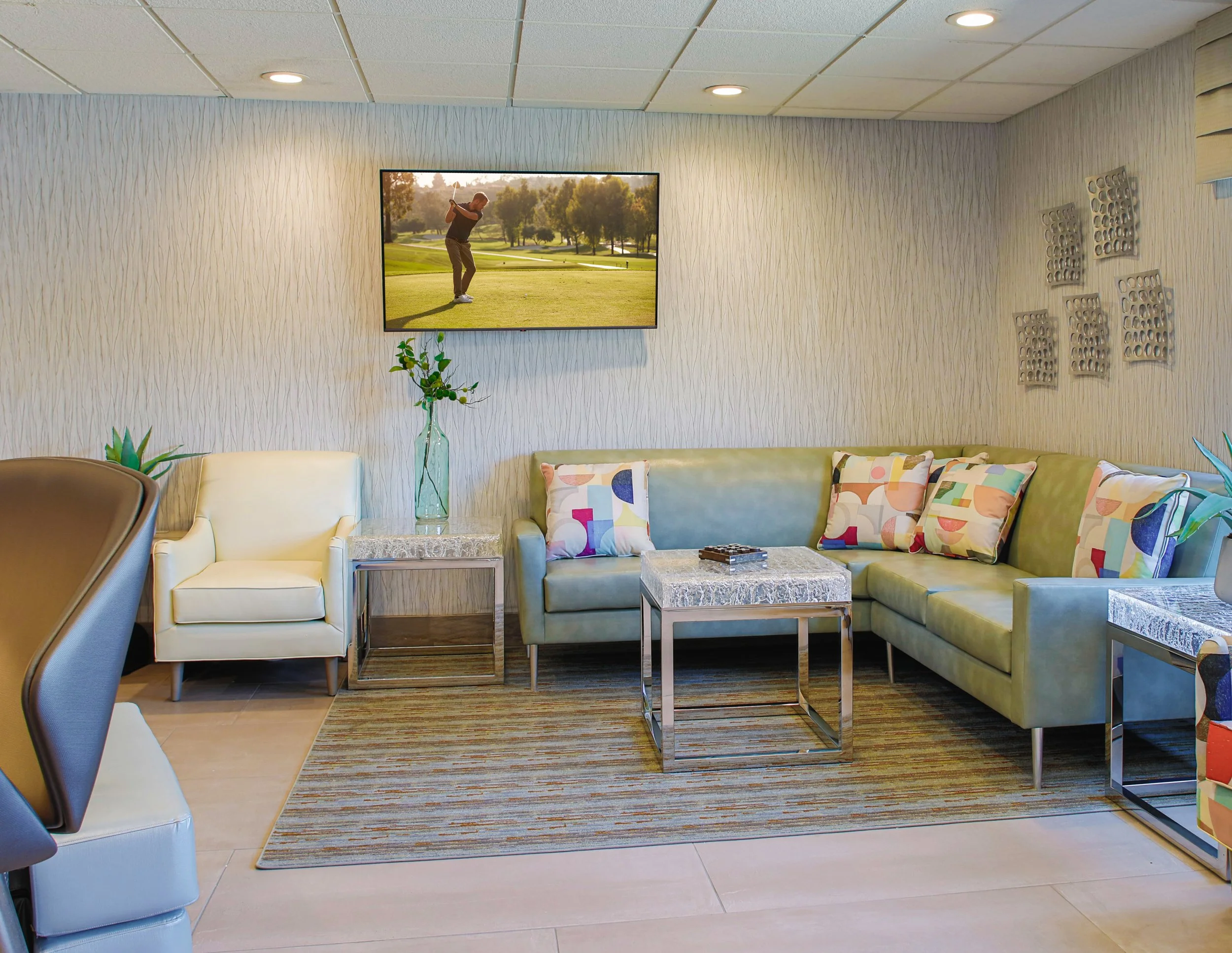 A modern living room with a green sectional sofa, colorful patterned pillows, a white armchair, a glass side table with a green plant, a marble-top coffee table, decorative wall art, and a flat-screen TV showing a golfer on a golf course.