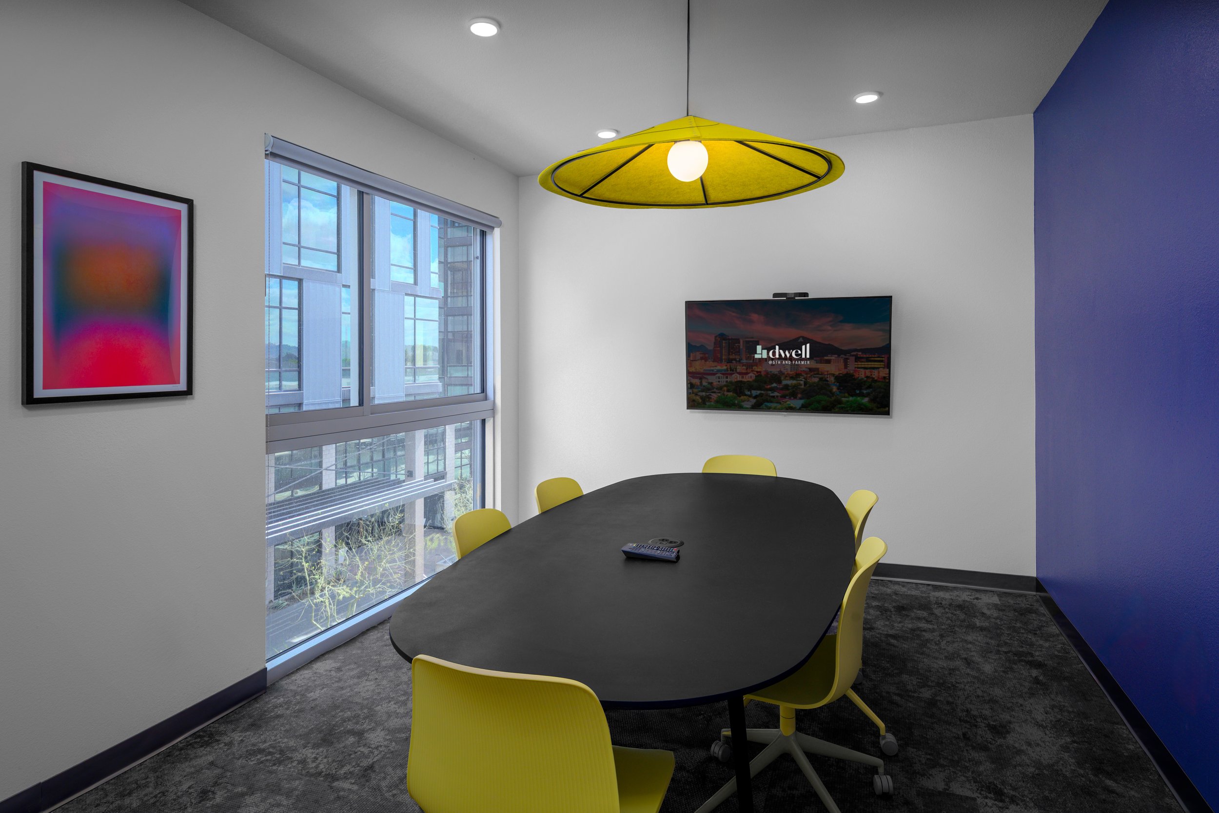 Modern conference room with a black oval table, yellow chairs, a large window with a city view, a flat-screen TV on the wall displaying "dwell," a colorful abstract artwork, and a yellow ceiling light.