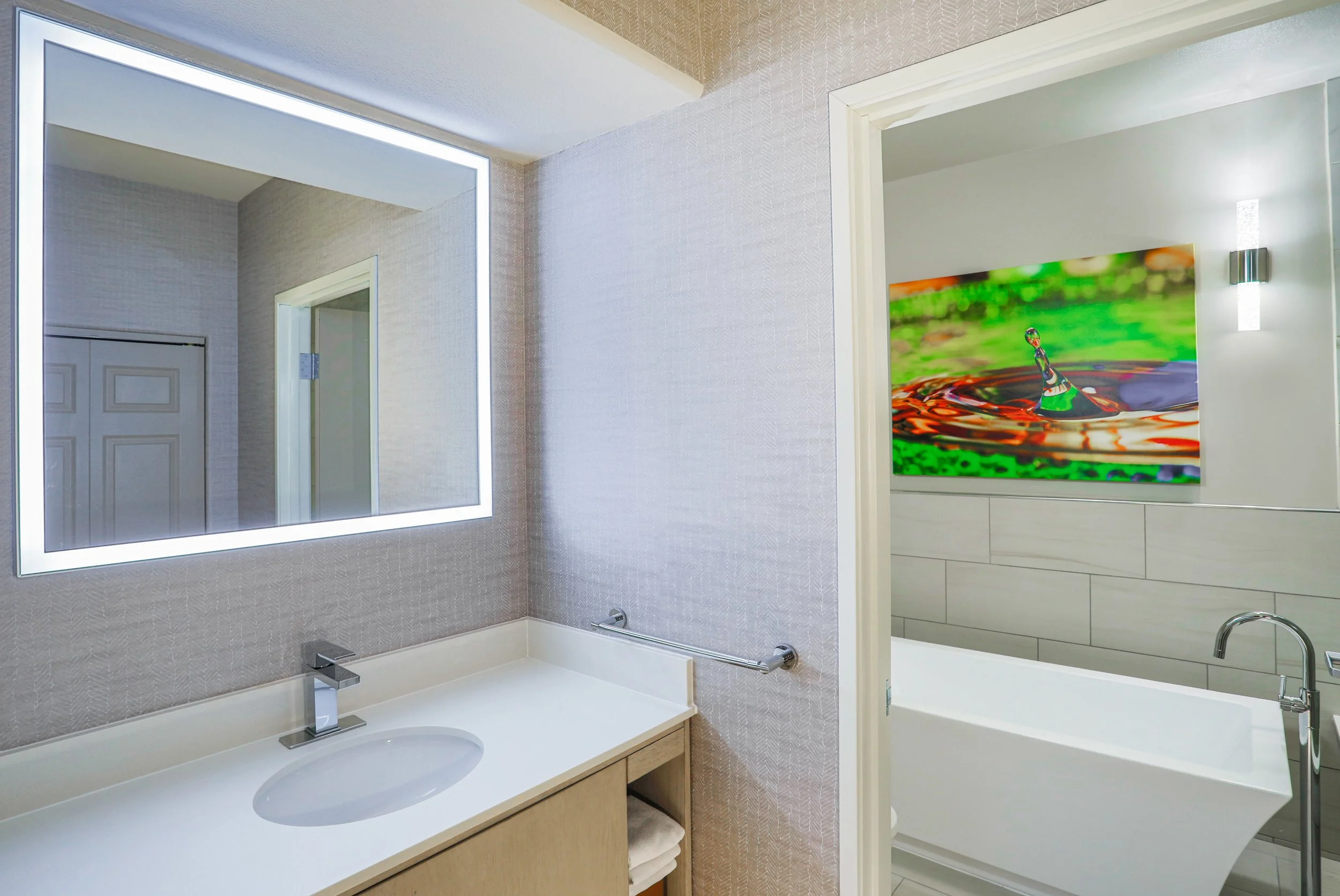 Modern bathroom with large illuminated mirror, beige vanity with towels, and a separate shower area with colorful artwork on the wall.
