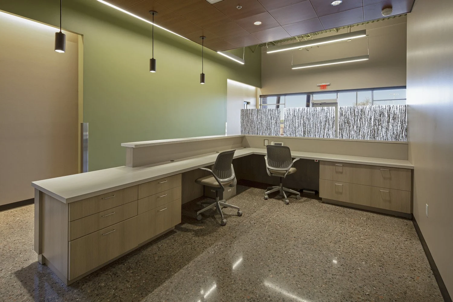 An empty office reception or front desk area with a white counter, two office chairs, and a large window with a decorative privacy screen, in a modern building.