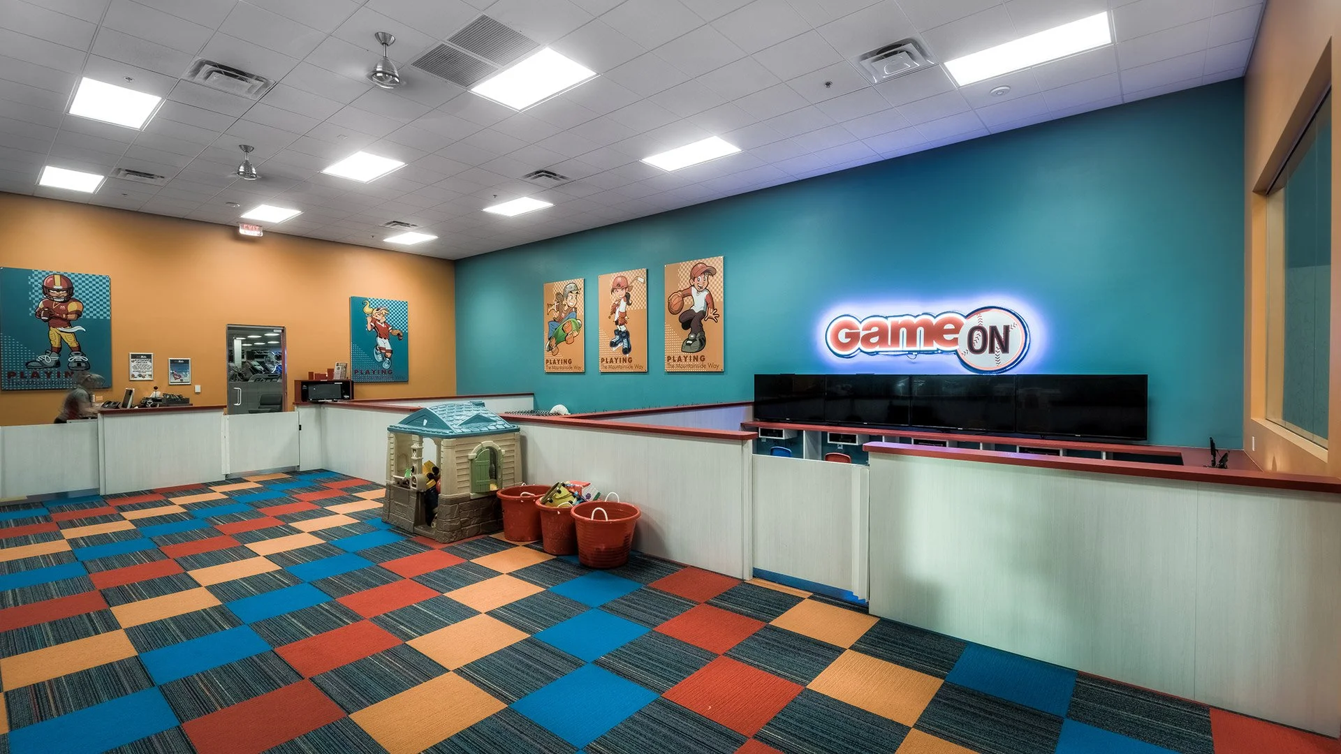An indoor arcade area with colorful checkered carpet, a white barrier enclosing a play area with toys, and a neon sign that says 'Game ON' on a teal wall, decorated with cartoon posters of children playing sports.