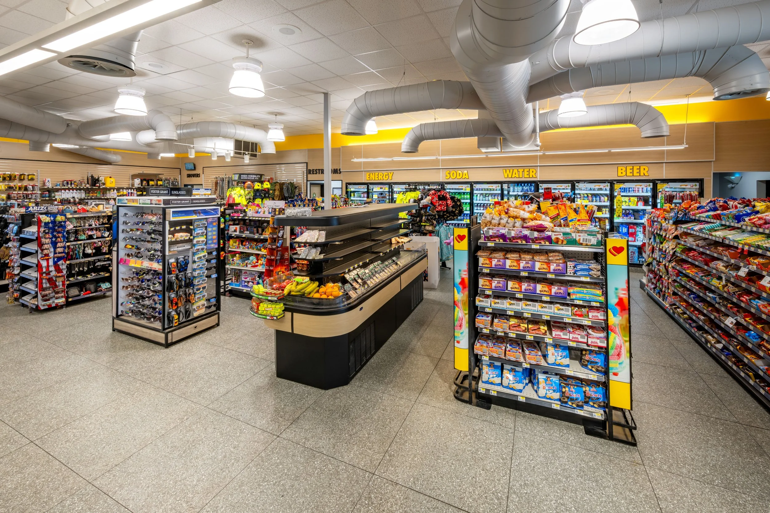Inside a convenience store with shelves stocked with snacks, drinks, and other items. Overhead signs indicate sections for energy drinks, soda, water, and beer.