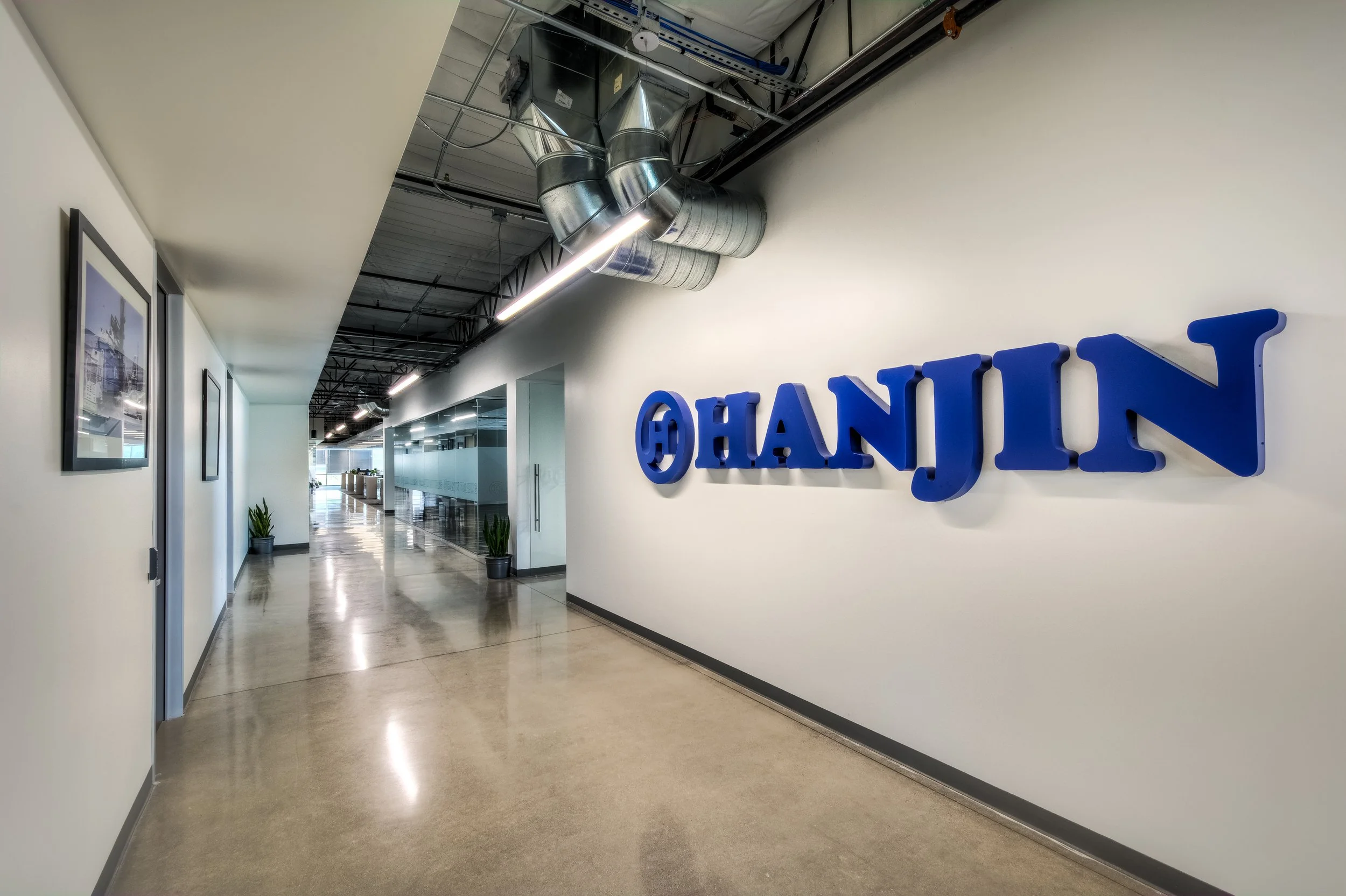 Interior of an office hallway with white walls, framed artwork, potted plants, and a glass-enclosed conference room. Blue 3D letters spelling 'Hanjin' are mounted on the wall. Exposed ceiling with ductwork and lighting.