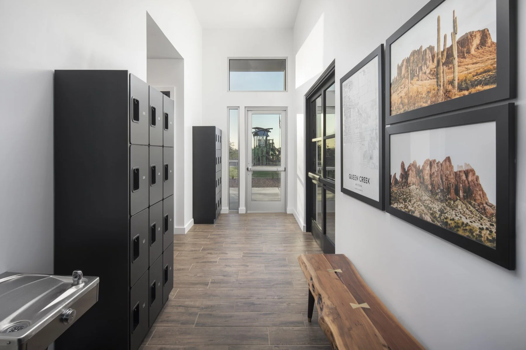 Locker room with black lockers, a wooden bench, framed landscape photos on white wall, and glass door leading outside.