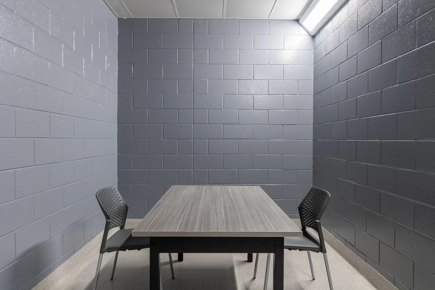 Small, empty interrogation room with gray painted cinder block walls, a light gray ceiling, and fluorescent lighting; contains a rectangular table and two black chairs with perforated backs.