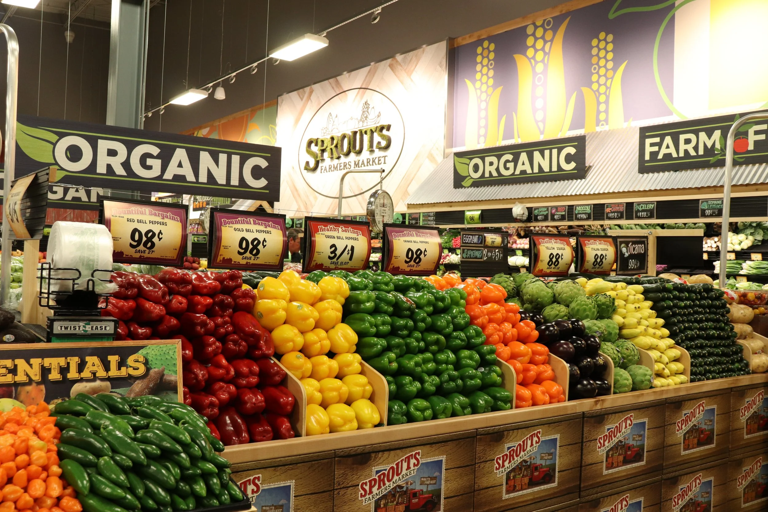 Display of various colorful vegetables arranged in sections at Sprouts Farmers Market, including red, yellow, green, orange, purple vegetables, with signs indicating prices and organic labels.